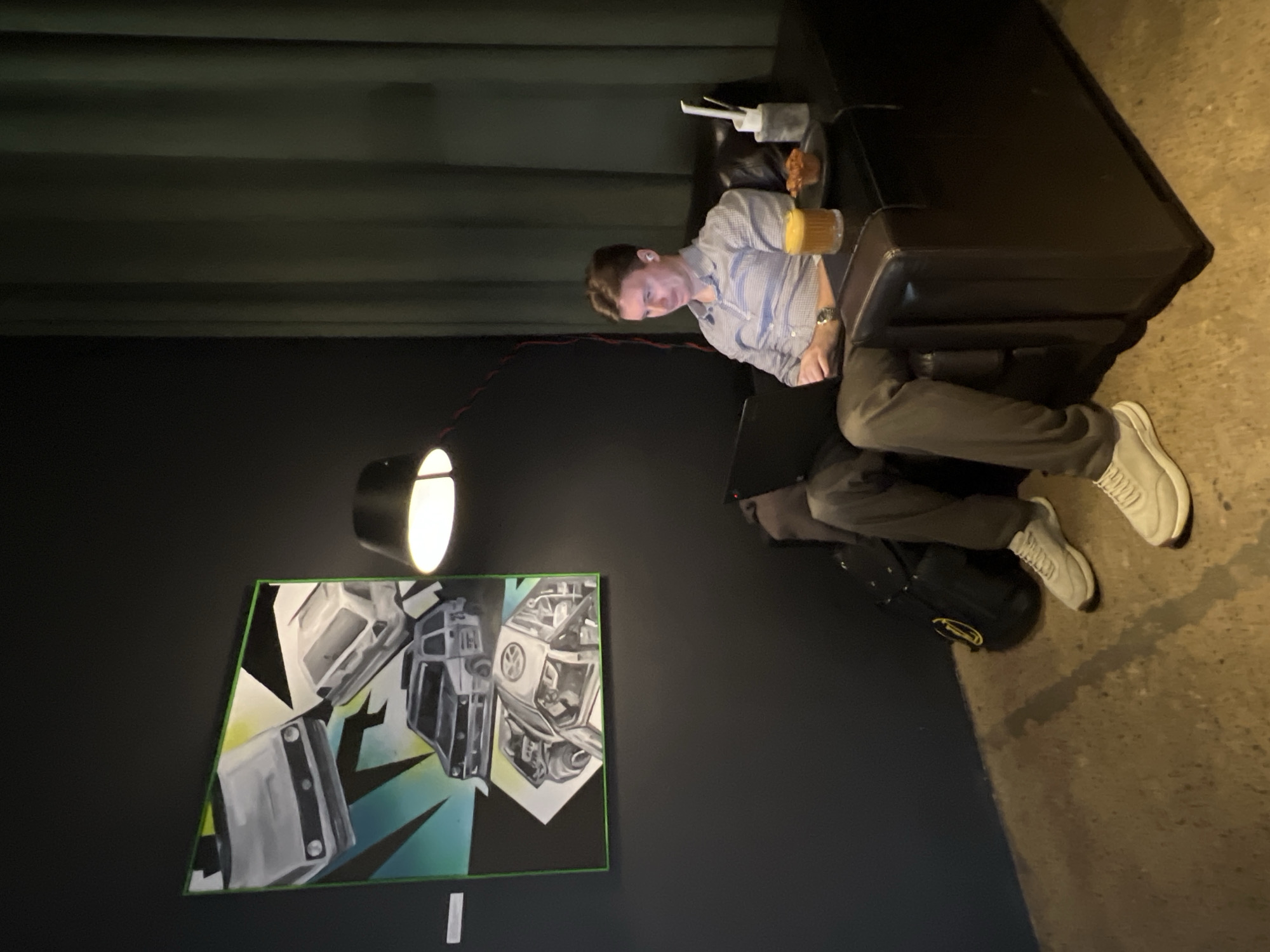 A man is seated at a dark leather couch or booth in what appears to be an indoor establishment. He is wearing a light-colored, button-down shirt, grey trousers, and white sneakers. He is holding a glass of amber liquid, possibly beer, in his right hand. On the dark surface to his right, there is a plate of food and a container of utensils.
Behind him, a black wall features a painting of what seems to be van models on a green and white backdrop. A spotlight is positioned above the artwork, illuminating it. The location might be Saint Petersburg, Russia, but this requires more information. There is a dark curtain in the upper left corner and a glimpse of a concrete-like floor on the right side. A dark bag is positioned near his feet.