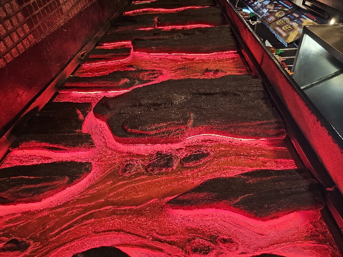 The image portrays the interior of a Dean & Dennys "Lava" themed fast-food restaurant, located in Buenos Aires, Argentina. The scene's dominant feature is a long, textured wall composed of dark, rugged panels with deep, organic grooves and fissures. These channels are intensely illuminated from within by bright red light, creating a vivid impression of glowing lava flows. Above this illuminated wall, a dark, flat counter displays at least three condiment bottles: two with red bodies and orange caps, consistent with Sriracha hot sauce, and one with a yellow cap. Behind the counter, a series of vertically arranged digital screens display menu items, including images of burgers labeled "Doble Cuarto de Libra," "Cheeseburger," "Bacon," "Papas Fritas" (French Fries), and "Milkshake." A dark, angular metal ventilation hood or ceiling element is situated above the menu screens. The floor in the foreground is dark, tiled with a subtle grid pattern, and reflects the ambient red light from the wall. No individuals are visible within the frame.