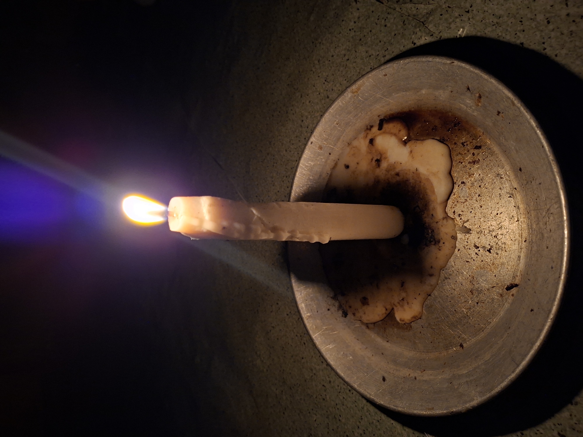 A lit candle sits horizontally in the center of a shallow, round, metallic dish with melted wax and debris. The background is dark and textured. The location is unknown, though the user asked for results as if the location was Lhokseumawe, Indonesia.
