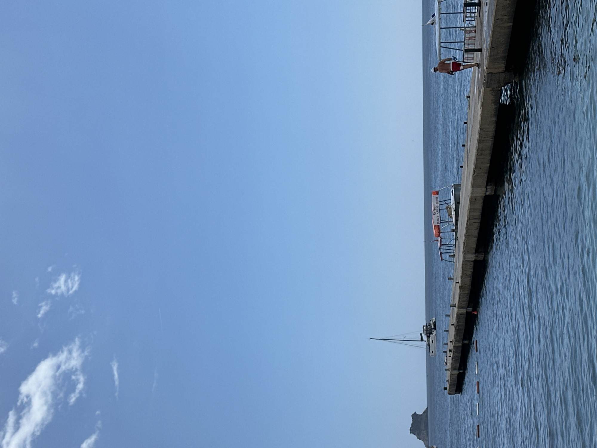 This media file captures a scene from a pier on a clear, sunny day, likely in the late morning or early afternoon, given the bright sky and sunlight on the water. The perspective is from the water looking towards the pier, which extends into the distance.
On the pier, a person is visible, wearing red shorts and a white cap, standing near the edge. The pier itself is made of concrete and appears to be a walkway over the calm, blue water. Several white and orange buoys are floating in the water near the pier, possibly marking a swimming area or a path.
In the background, a sailboat with its mast extended is anchored in the water. The sea stretches out to the horizon under a clear blue sky with a few wispy white clouds. A mountainous landmass is visible in the far distance on the left side of the image.
There is some visible text on a white rectangular object attached to the pier, which reads "BUOY HAWAI" and "BUOY". This, along with the mention of Budva, Montenegro in the location context, suggests the location is a coastal area, likely a beach or harbor in Budva. The overall mood of the image is serene and tranquil, depicting a typical summer day at the seaside.