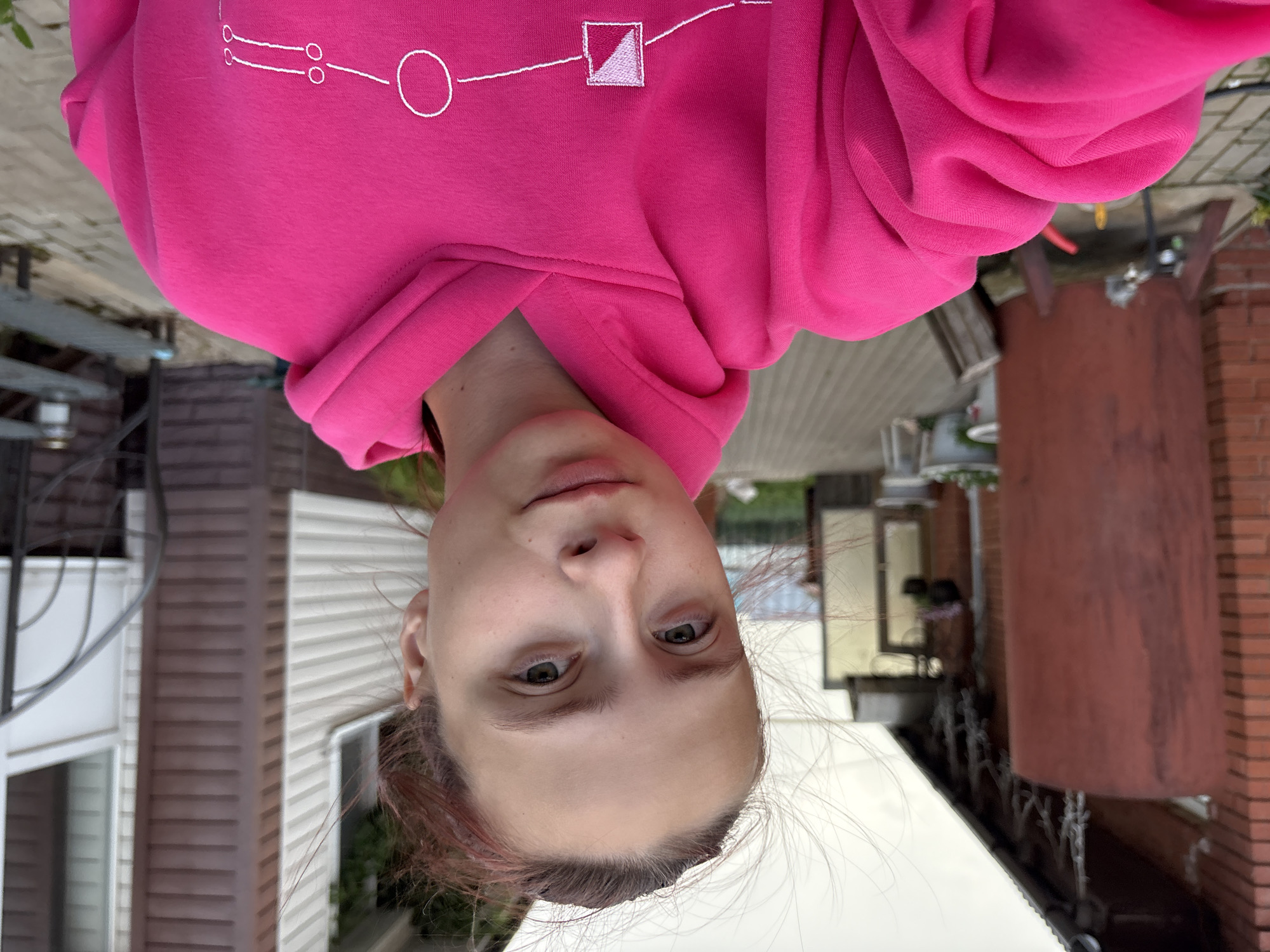 A young woman with reddish-brown hair pulled back and wisps framing her face, and green eyes, is taking a selfie outdoors. She is wearing a bright pink hoodie with a white embroidered design on the chest. Behind her, to the left, is a brick building with a dark red cylindrical object in front of it. To the right is a building with light-colored siding and a white door, next to a dark metal spiral staircase. The ground is paved with grey bricks. The background suggests a semi-rural or suburban environment. The overall lighting indicates an overcast day.