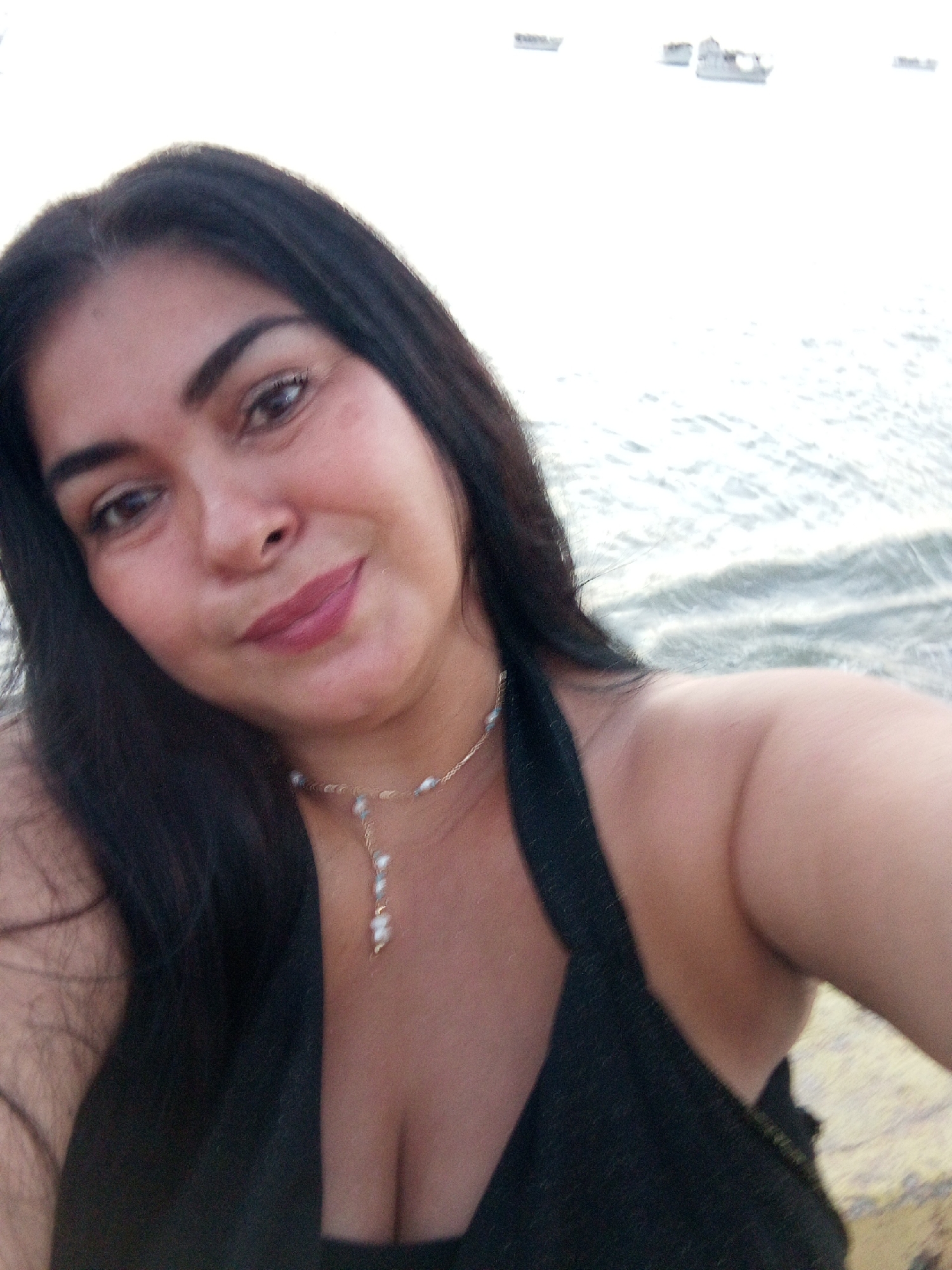 A medium shot shows a dark-haired woman posing outdoors, possibly on a beach or near a body of water. Her dark hair contrasts with the lighter backdrop, which consists of water and possibly boats or structures in the distance. She wears a dark top and a light-colored necklace. The background suggests a beach or coastal setting, which aligns with Juan Griego, Venezuela, known for its beaches and coastal areas.