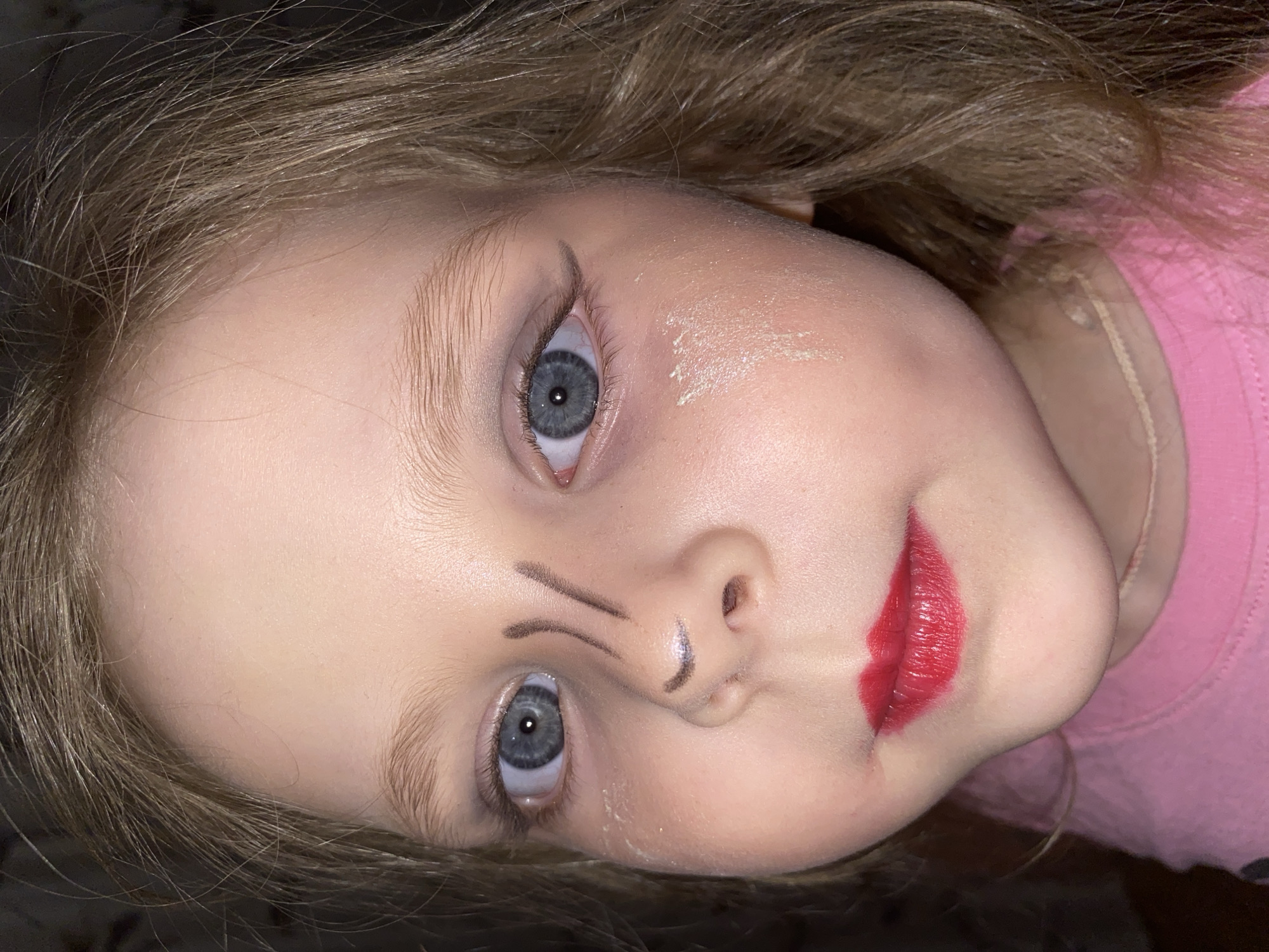 This close-up portrait, taken in Votkinsk, Russia, features a young child, likely a girl, with fair skin, light blonde or light brown hair, and striking blue-grey eyes. The image is rotated, presenting the child's head at a significant tilt, with her gaze directed towards the viewer.
The child's face is adorned with what appears to be play makeup or a costume application. Her lips are coated in a vibrant, glossy red lipstick, applied with an exaggerated, almost doll-like shape. Above her natural eyebrows, bold, arched dark lines are drawn to create a theatrical eyebrow effect. A small dark line is also visible on the bridge of her nose, and another short line on the tip. On her left cheek (viewer's right) and near her forehead, there are faint streaks of a light, possibly iridescent or glittery substance, resembling white paint or shimmer.
Her hair, a mix of light brown and blonde strands, frames her face. She is wearing a pink, ribbed top, visible on the right side of the frame. The child's expression is neutral to slightly serious, with wide, open eyes that appear curious or focused. The lighting suggests an indoor setting. The image captures a moment of imaginative play or dress-up. No visible text is present in the media file.