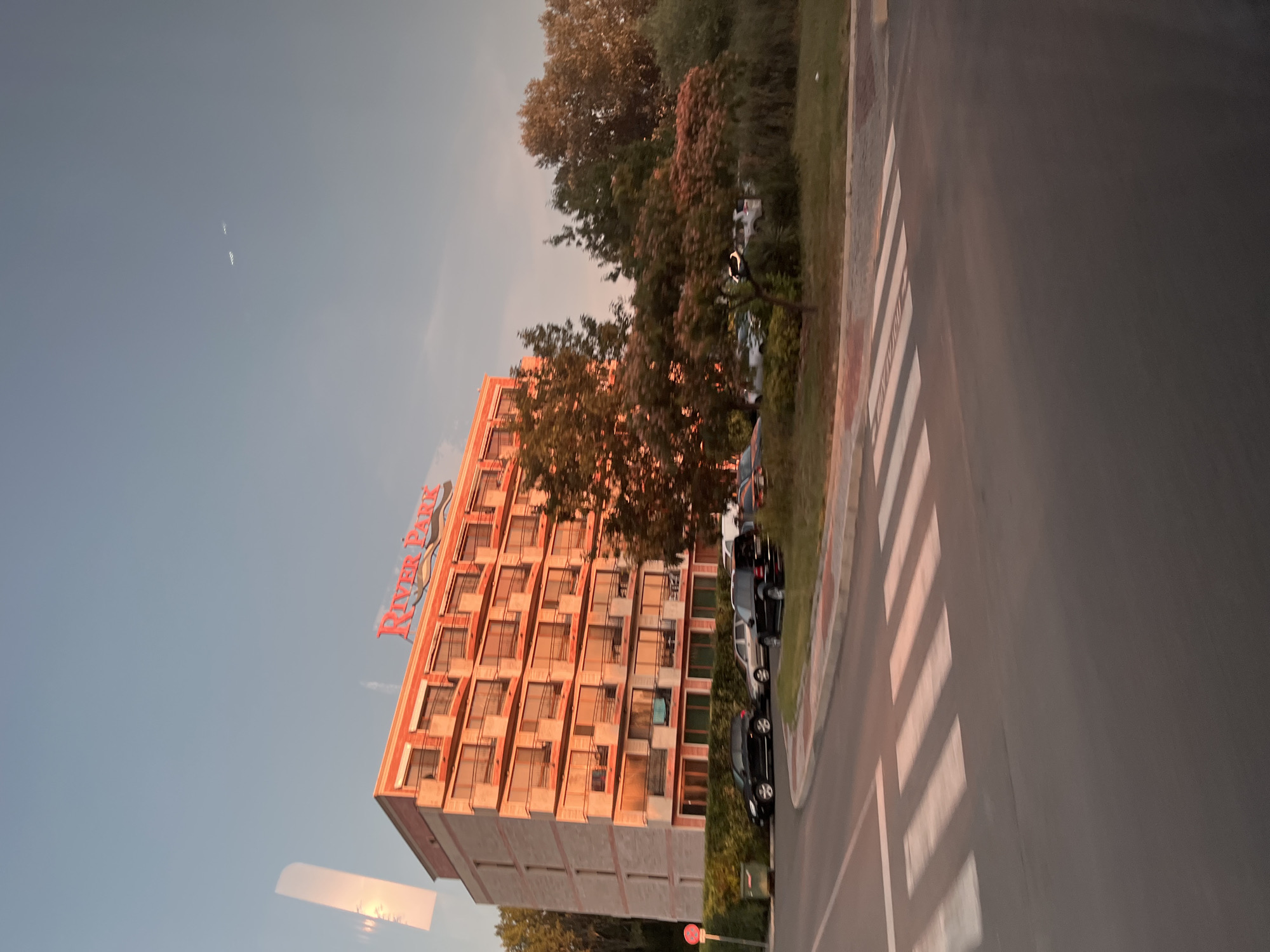 A building with the "River Park" sign is visible in the city of Nesebar, Bulgaria. The building is tall and has multiple floors with balconies. The sun is setting, casting a warm glow on the building and trees. Cars are parked in front of the building, and a pedestrian crossing is visible on the road. The sky is clear with a few distant airplanes.