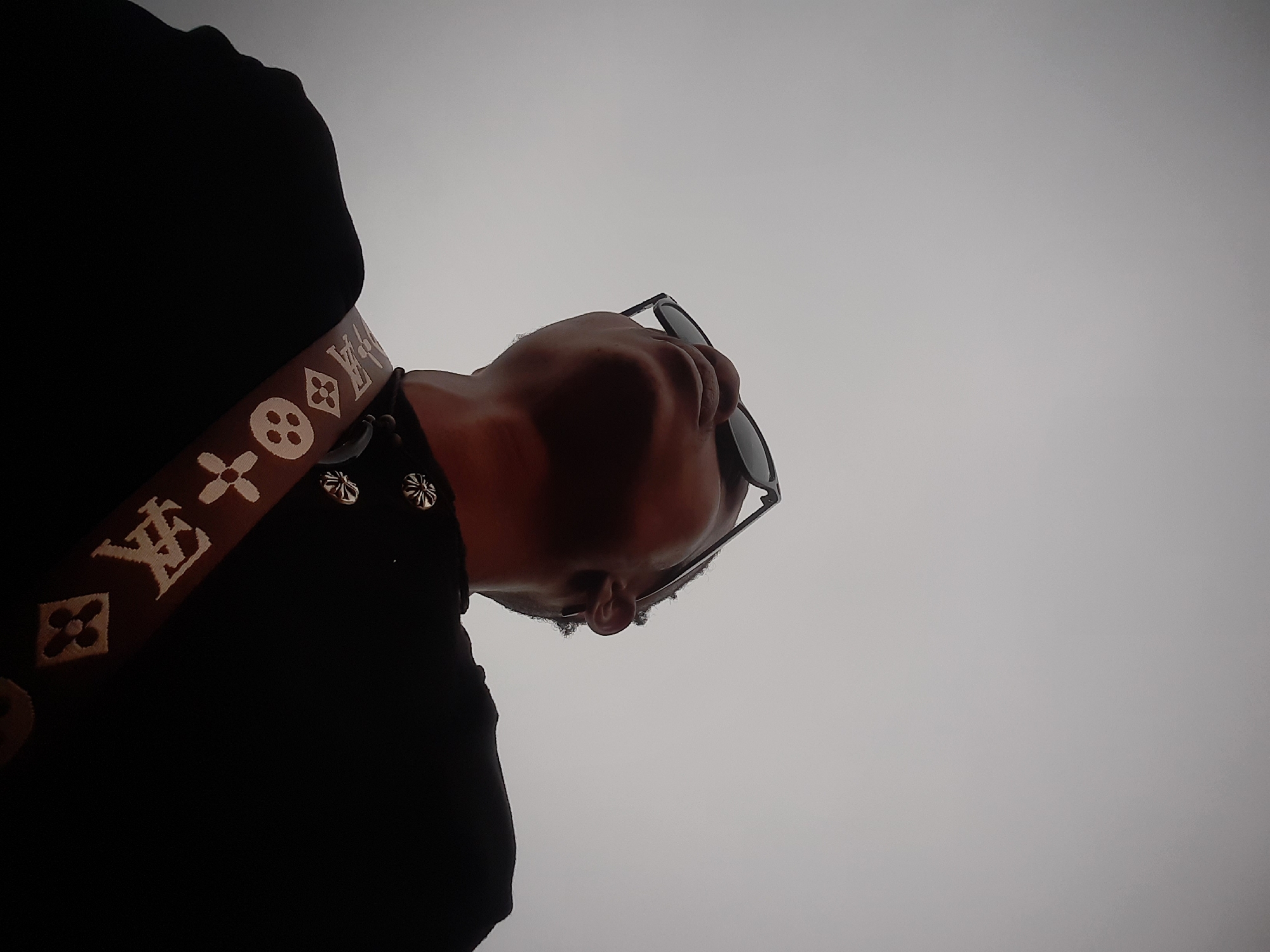 A low-angle shot depicts a person wearing sunglasses and a dark-colored shirt or top, with a brown patterned strap or belt featuring logos and metallic details. The background is a bright, neutral sky or overcast sky. The subject appears to be looking down. It is not possible to ascertain the location of the photo in Jos, Nigeria based on the visual information.