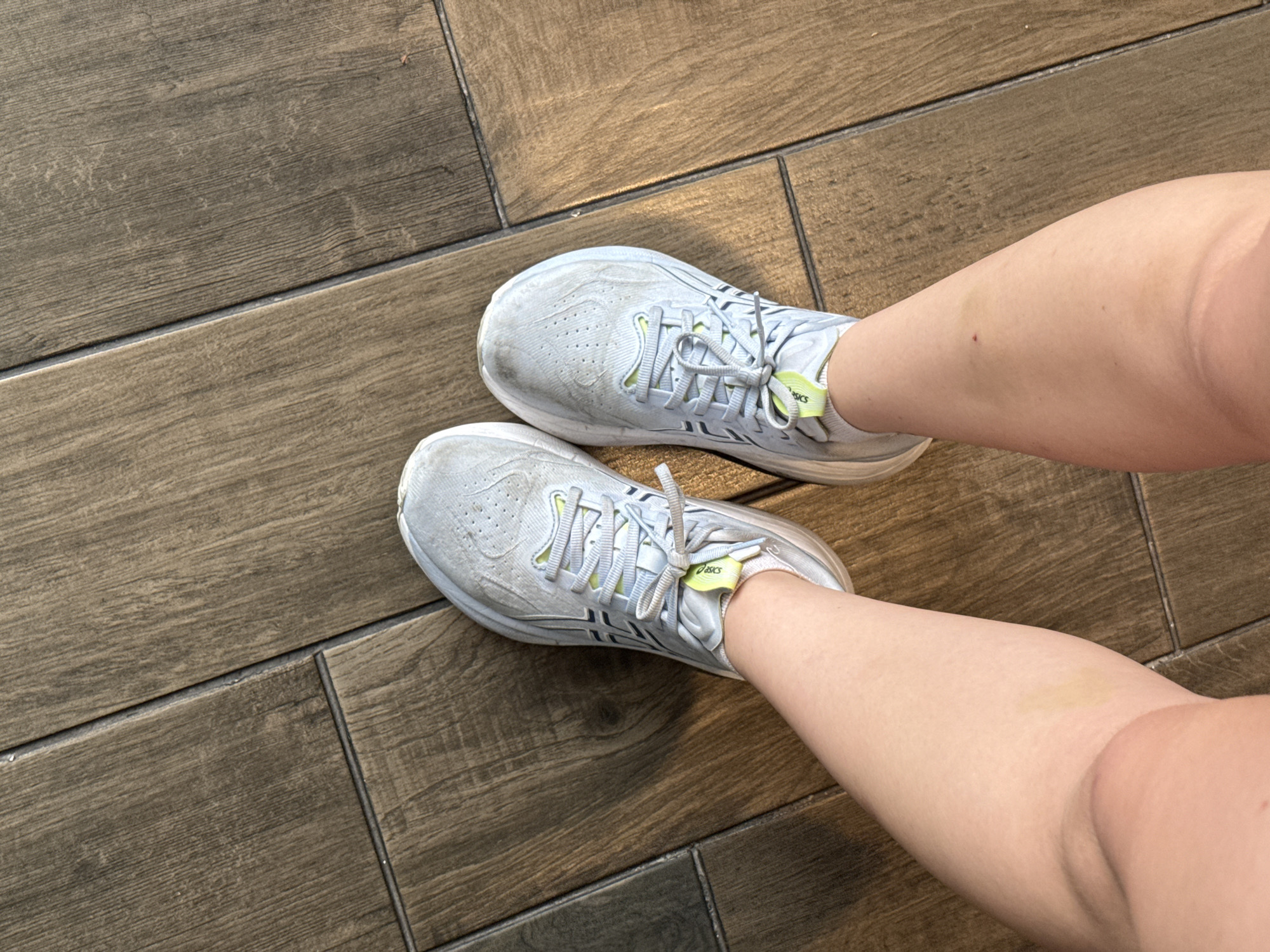 The image shows a view from above of a person's legs and feet wearing white Asics running shoes. The shoes appear to be used, with dirt and wear visible on the outer surface. The legs are pale in color and have some visible bruising. The shoes and legs are positioned on a floor made of dark, wood-look tiles. The photograph was taken indoors. There is no information to confirm this image was taken in Mexico City, Mexico.