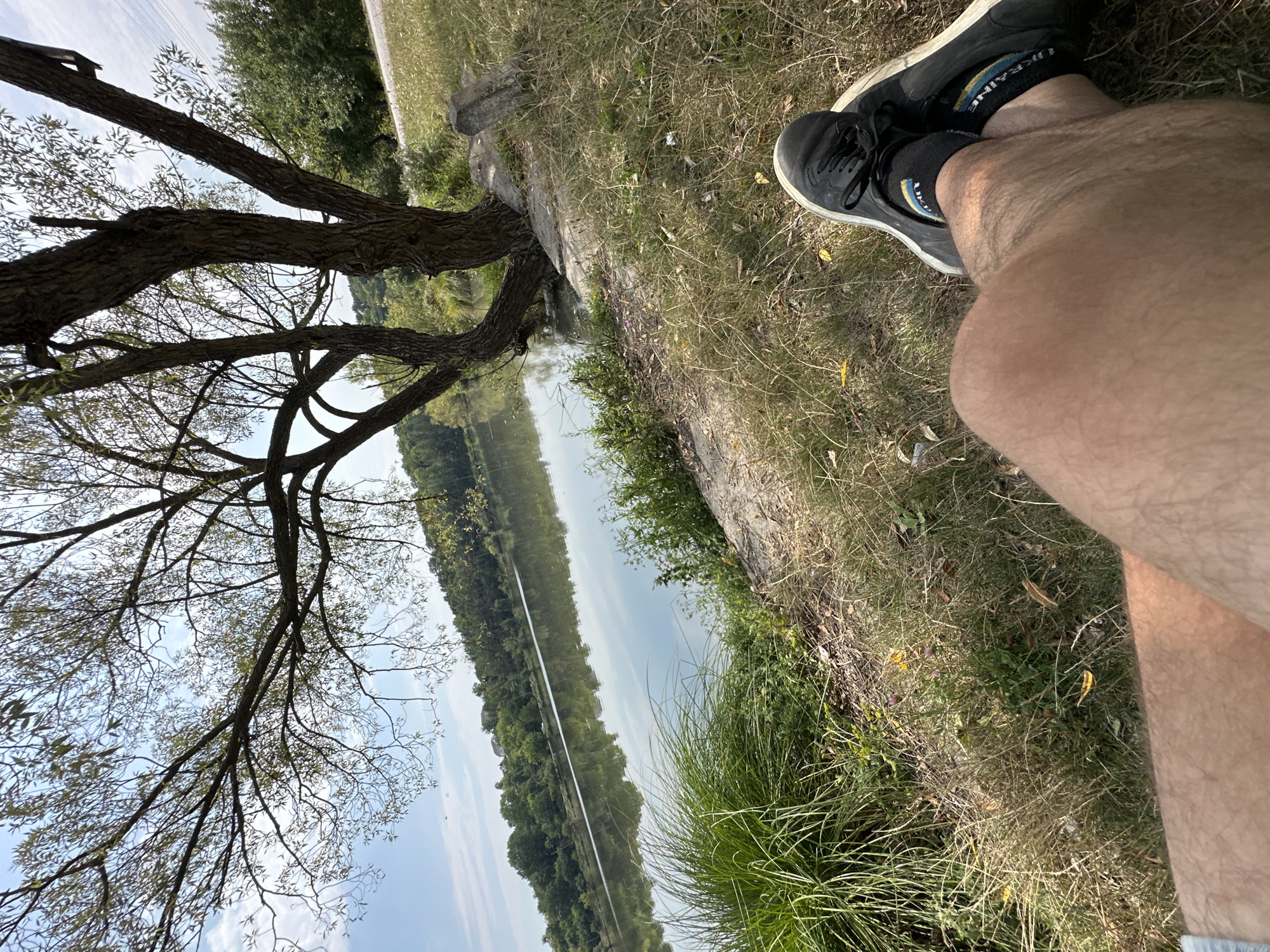 The image depicts a relaxed scene by a body of water, likely a river or lake, with a person's legs in the foreground. The person is wearing black sneakers and socks with yellow and blue stripes, suggesting a connection to Ukraine, as the colors are similar to the Ukrainian flag. They are sitting on dry grass, with a large tree to their left, its branches extending towards the sky. The water reflects the surrounding greenery and the blue sky with scattered clouds. In the background, a lush green treeline lines the opposite bank, with some buildings faintly visible on a hill. The overall setting appears to be a natural, possibly park-like area. While the image does not provide explicit confirmation of the location as Makiv, Ukraine, the footwear's colors may hint at it. The scene is peaceful and suggests a moment of quiet contemplation or enjoyment of nature.