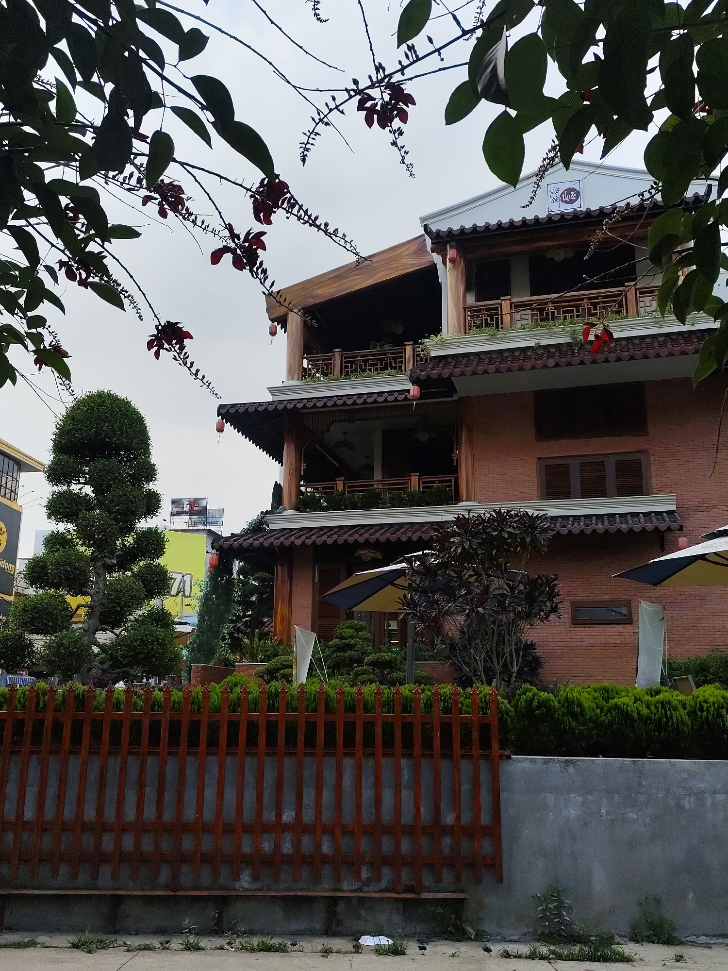 This image captures a multi-story building with distinctive traditional Vietnamese architecture, possibly a café or restaurant, under an overcast sky in Da Lat, Vietnam. The building is constructed with reddish-brown bricks and features dark, curved tiled roofs with upturned eaves. Multiple wooden balconies with ornate railings and exposed wooden pillars are visible, suggesting outdoor seating areas. Red lanterns hang decoratively from some eaves.
The foreground is framed by dark green leaves and branches adorned with small, red, pendulous flowers. Below the building, a meticulously maintained garden features a tall, sculpted topiary tree with multiple tiers on the left, and neat hedges of green shrubs along a reddish-brown wooden picket fence. Beyond the fence, a grey concrete wall and a sidewalk with sparse weeds are visible. Two large outdoor umbrellas, one with yellow and black stripes, are positioned near the building, indicating a patio or garden seating area.
In the mid-ground, a white rectangular sign on the uppermost part of the building clearly displays the stylized text "Việt Thảo" accompanied by a graphic element resembling a sprig or branch, suggesting the name of the establishment. To the far left, parts of other buildings are visible, including a yellow structure with an indistinct sign and another distant building with a red and white sign on its roof, though the text on these is unreadable. No people or specific activities are depicted, giving the scene a tranquil, static quality. The soft, even lighting indicates a cloudy day, possibly morning or afternoon.