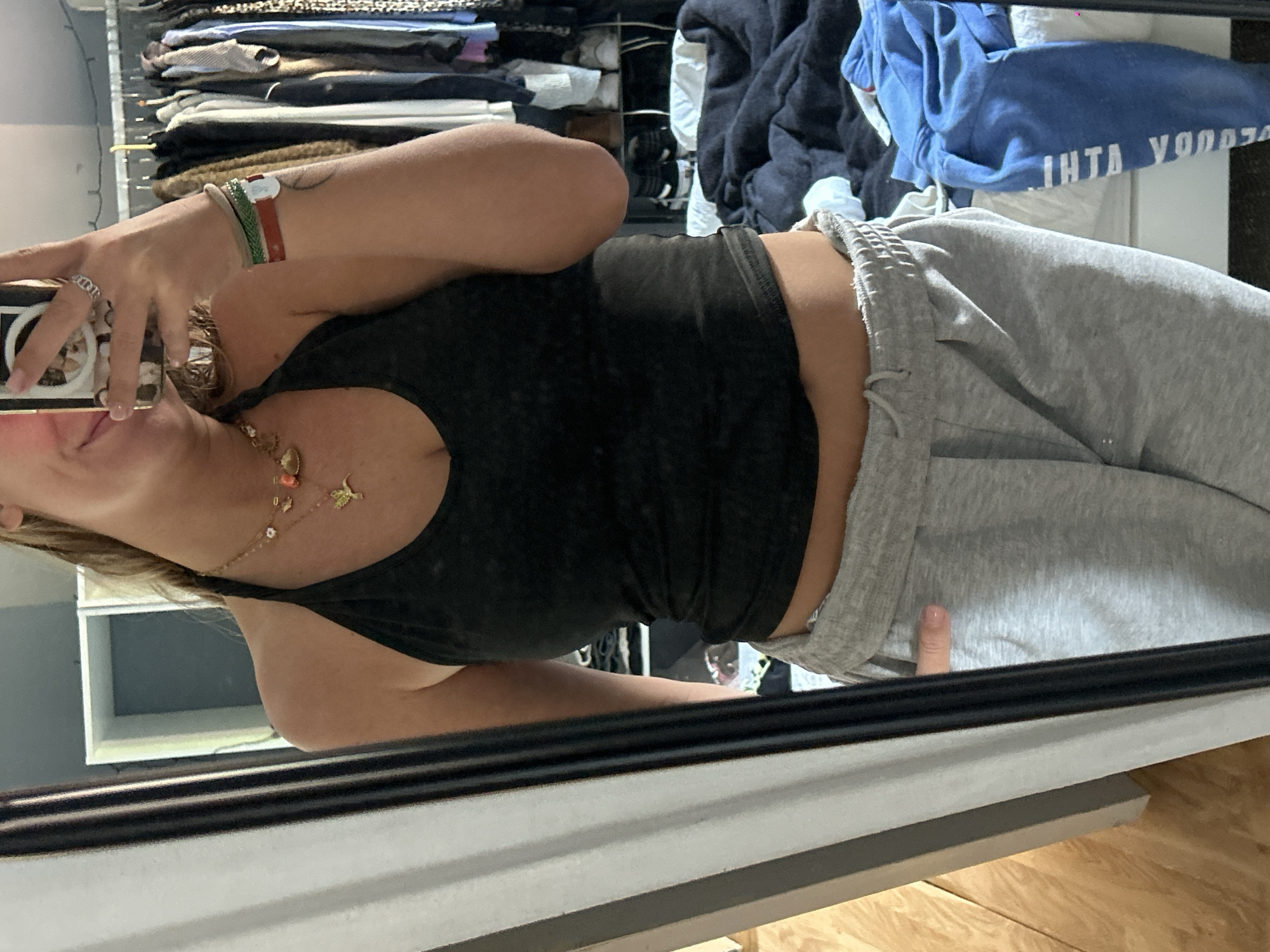 A mirror selfie, taken in Lannoy, France, depicts a person from the chest down to the upper thighs, with the image rotated 90 degrees clockwise. The subject is wearing a black tank top and light grey sweatpants, with a thumb tucked into the waistband. Their right hand holds a smartphone, capturing the image, and partially obscures the lower part of their face from the nose down.
On the subject's right wrist are multiple bracelets, including green, red, and light-colored bands. A silver-toned ring is visible on the right ring finger. A delicate gold-colored necklace adorns the neck, featuring several small charms such as a coral-colored bead, a golden bird or insect, a crescent moon, and white flower-like elements.
The background, reflected in the mirror, reveals an indoor setting, possibly a bedroom or dressing area, with shelves stacked with folded clothes. Among these, a blue hoodie or sweatshirt is visible, bearing reversed and partially obscured text which appears to read "IHTA YOO", possibly "ATHI" when corrected for reflection. The overall scene suggests a casual and private moment.
