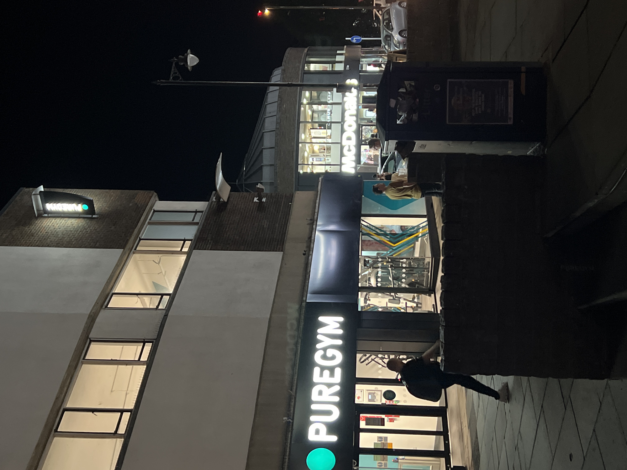 The image shows a nighttime street scene in Guildford, United Kingdom. On the left, a PureGym is illuminated with its prominent sign. A person wearing a dark backpack is walking away from the gym. To the right, a McDonald's restaurant with a rounded glass facade is visible, also lit up. Several cars are parked in front of McDonald's. A lamppost stands on the right side of the street. A lit traffic signal with a green light is mounted on the brick building to the left. A dark-colored trash receptacle is situated in the foreground between the gym and McDonald's. The ground is paved with square tiles.