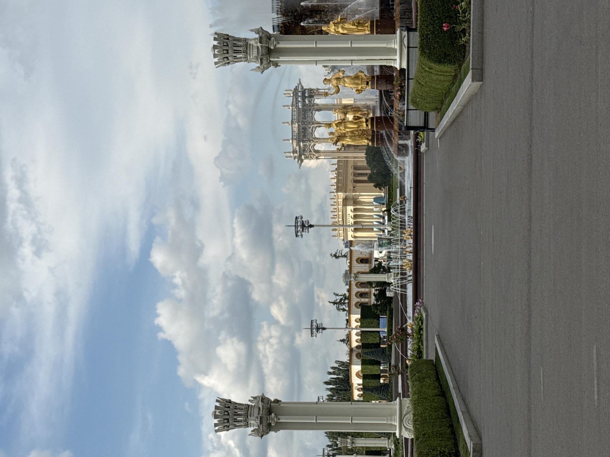The image shows a view of the Friendship of Peoples fountain at VDNKh park in Moscow, Russia. The fountain features golden statues of women representing different Soviet republics, set against a backdrop of classical architecture and lush greenery. The water jets of the fountain are visible, creating a dynamic and majestic scene. The sky is partly cloudy, with a mix of blue sky and white cumulus clouds. The foreground shows a paved pathway leading towards the fountain, with neatly trimmed hedges lining the sides. The overall impression is one of grandeur and artistic achievement, typical of the Soviet era's architectural style.