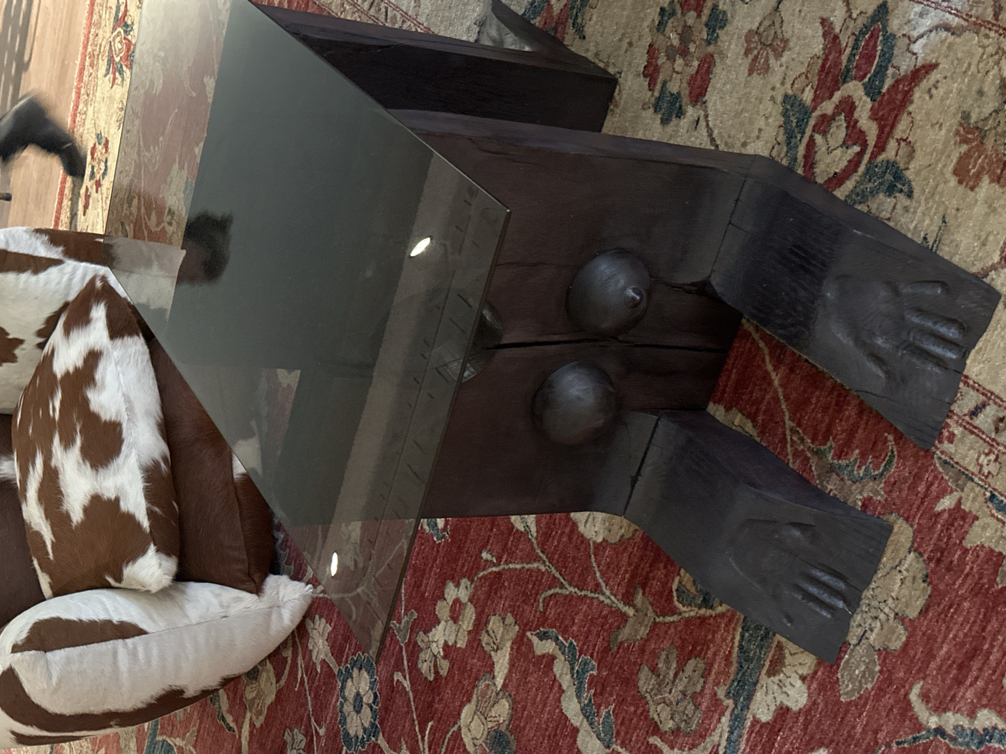 The image shows a section of a room with a dark wooden coffee table featuring a glass top. The table appears to be uniquely designed with sculpted details, possibly resembling feet or hands at the ends. On the left side of the table, there is a patterned cushion or pillow with brown and white patches. The table and cushion are placed on a large rug with a red and beige floral pattern. The location in London, United Kingdom, is not evident in the image.