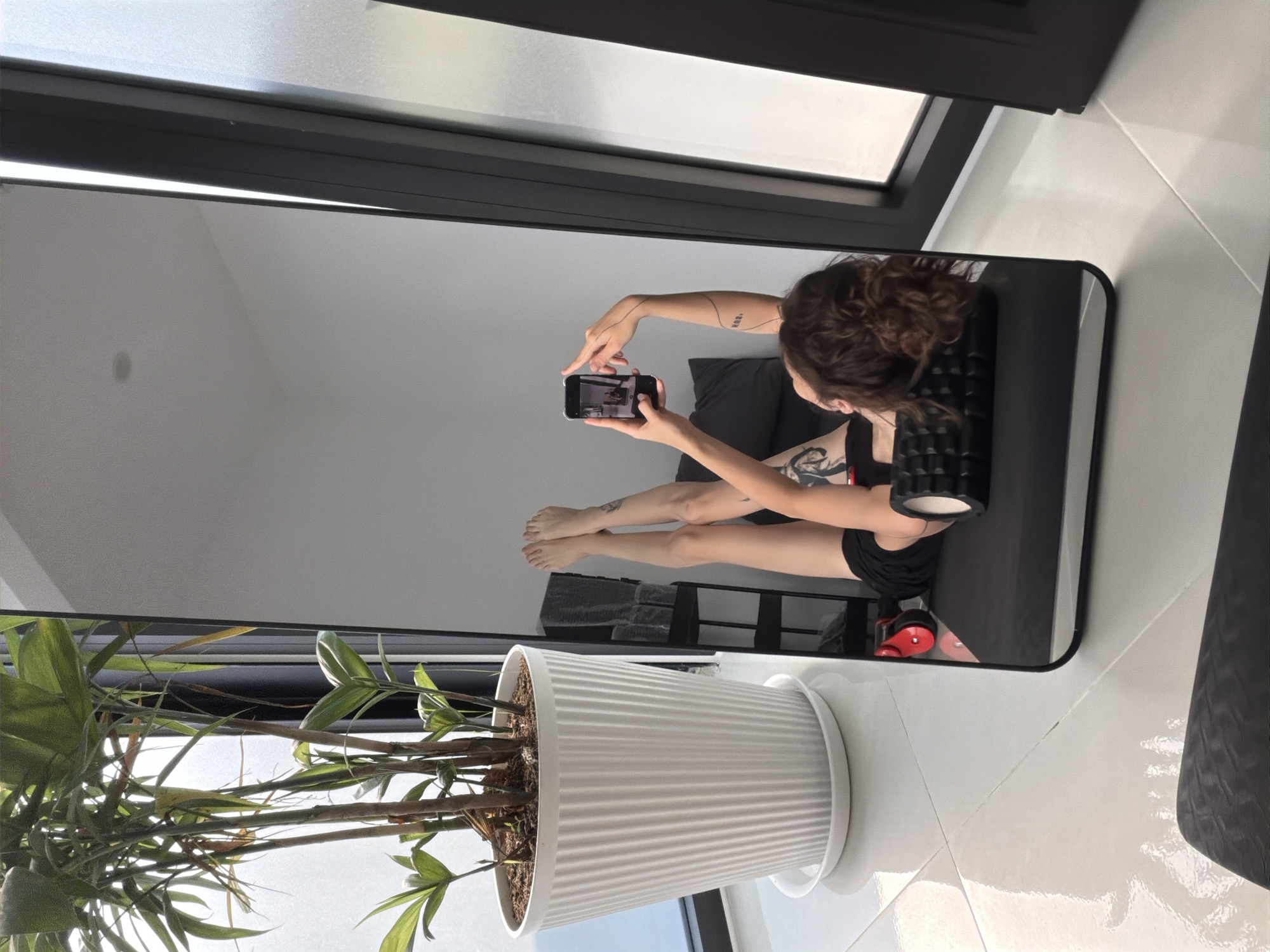The image shows a person's reflection in a large, black-framed mirror. The person is holding a smartphone and appears to be taking a picture of themselves, while sitting on a dark surface and leaning against a textured, cylindrical object. The person has curly brown hair, tattoos on their arms, and is wearing dark clothing. The room appears to be well-lit with white walls. In the foreground, a large potted plant with green leaves is visible. The background also shows a window and other furniture. The exact location in Đà Nẵng, Vietnam cannot be determined from this image.