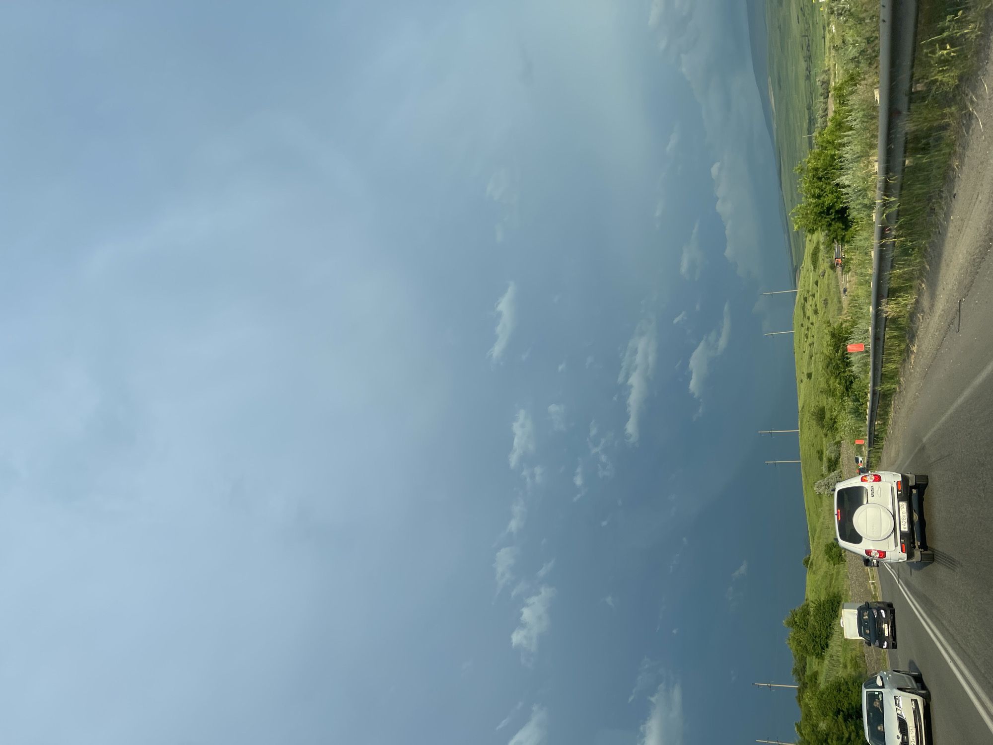 The image shows a view from inside a vehicle driving on a highway. The sky is overcast with dark, stormy clouds, particularly on the left side of the frame, suggesting impending rain. The road is paved, with multiple lanes visible. Several vehicles are on the road, including a white SUV in the foreground, a white sedan to its left, and a black SUV further ahead. The landscape on either side of the road consists of rolling green hills covered in grass and some trees. Power lines and utility poles are visible along the roadside. In the distance, hills stretch towards the horizon. The overall impression is a scenic drive under a dramatic sky, possibly near the city of Tatarka, Russia, given the context provided.
