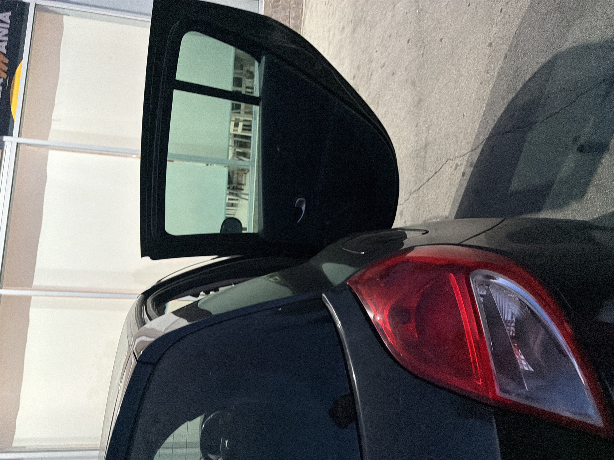 The image shows the rear quarter panel of a dark-colored car, possibly a hatchback, with the rear passenger door open. The car is parked on an asphalt surface with some visible cracks. In the background, there is a building with large, frosted windows, suggesting it might be a commercial establishment. A portion of a yellow and orange sign with text is visible at the top right of the frame, though the full text is not decipherable. The lighting indicates it is daytime, with shadows cast on the ground. The overall scene appears to be an outdoor setting in a city, potentially in Orhei, Moldova, based on the provided context. There are no people visible in the image.