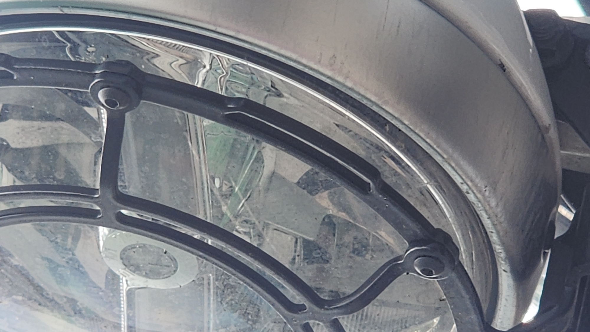 A close-up shot shows a round headlight with a black metal grid guard attached to a fender, likely part of a vehicle, possibly a motorcycle or car. The headlight is slightly dirty, and the fender has a metallic sheen. The background is indistinct. The location is not discernible from the image. There are no people visible.