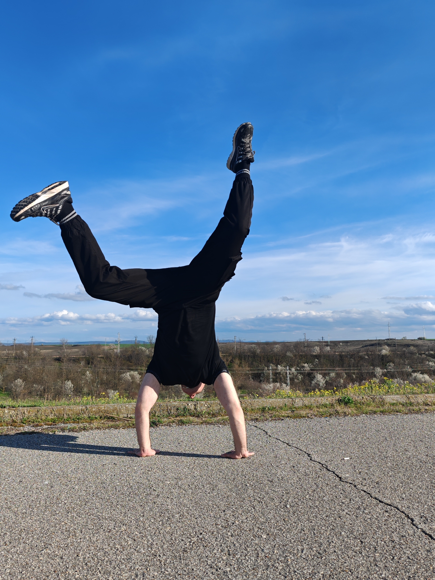 A man is performing a handstand on an asphalt road, presumably on the outskirts of Shumen, Bulgaria. The man is wearing black pants, a black t-shirt, and black and white sneakers. His legs are spread in a "V" shape against a blue sky with wispy clouds. The asphalt has a visible crack. Beyond the road is a grassy verge, followed by a vista of fields and trees in the distance. Overhead power lines are also visible in the background. The scene suggests an outdoor setting in a rural or semi-rural area near Shumen.