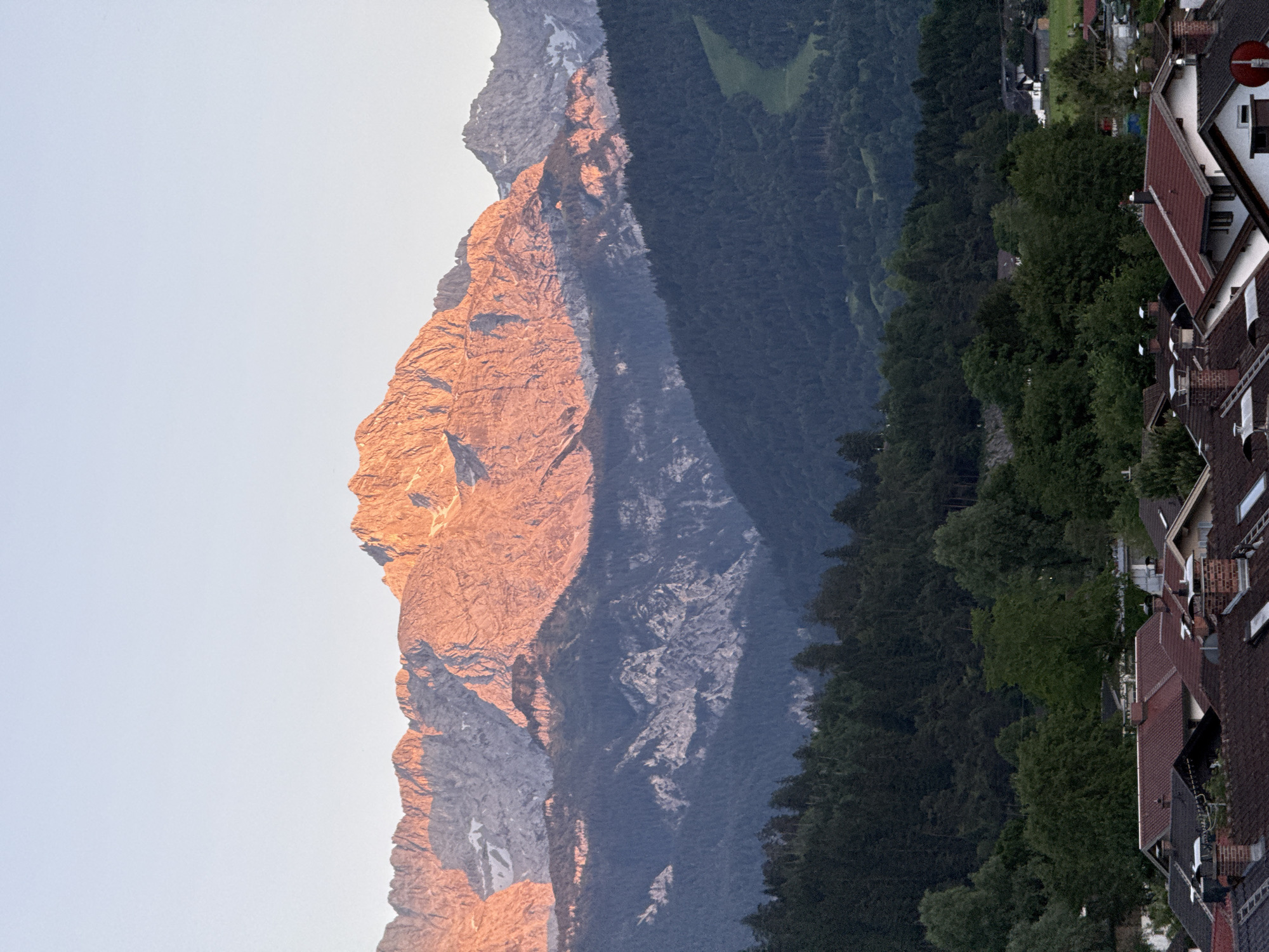 The image captures a mountain range, likely the Alps, bathed in the warm glow of sunrise or sunset. The prominent peaks are illuminated with hues of orange and pink, contrasting with the cooler, shadowed slopes and dark green forests in the midground. In the foreground, a village with red-tiled roofs and chimneys is visible, nestled at the base of the mountains. The sky above is a pale blue, gradually fading to a lighter shade near the horizon. The overall scene depicts a serene mountain landscape with human settlement at its foot, typical of locations like Garmisch-Partenkirchen, Germany, a town known for its alpine setting. No people or specific actions are depicted.