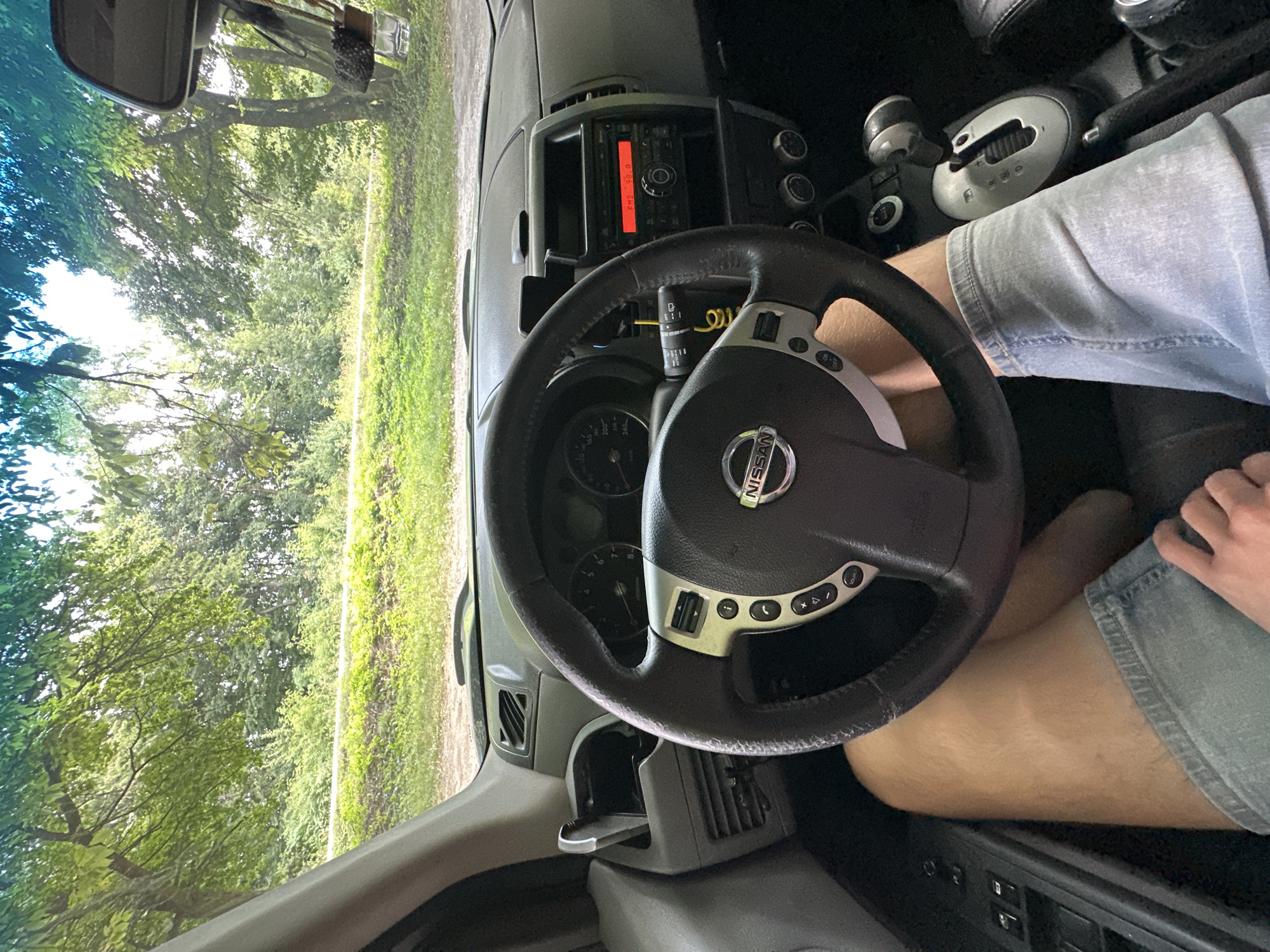 The image shows the interior of a Nissan car. The viewpoint is from the driver's seat, looking forward. The steering wheel is prominent in the foreground, with the Nissan logo visible. The dashboard contains a radio with a red display, a gear shift, and various buttons and vents. Outside the car, through the windshield, is a view of trees and greenery, suggesting a natural or rural setting. There are no people clearly visible in the image, though a person's legs wearing denim shorts are partially in view. The image does not provide any specific information about the location being Shatava, Ukraine.