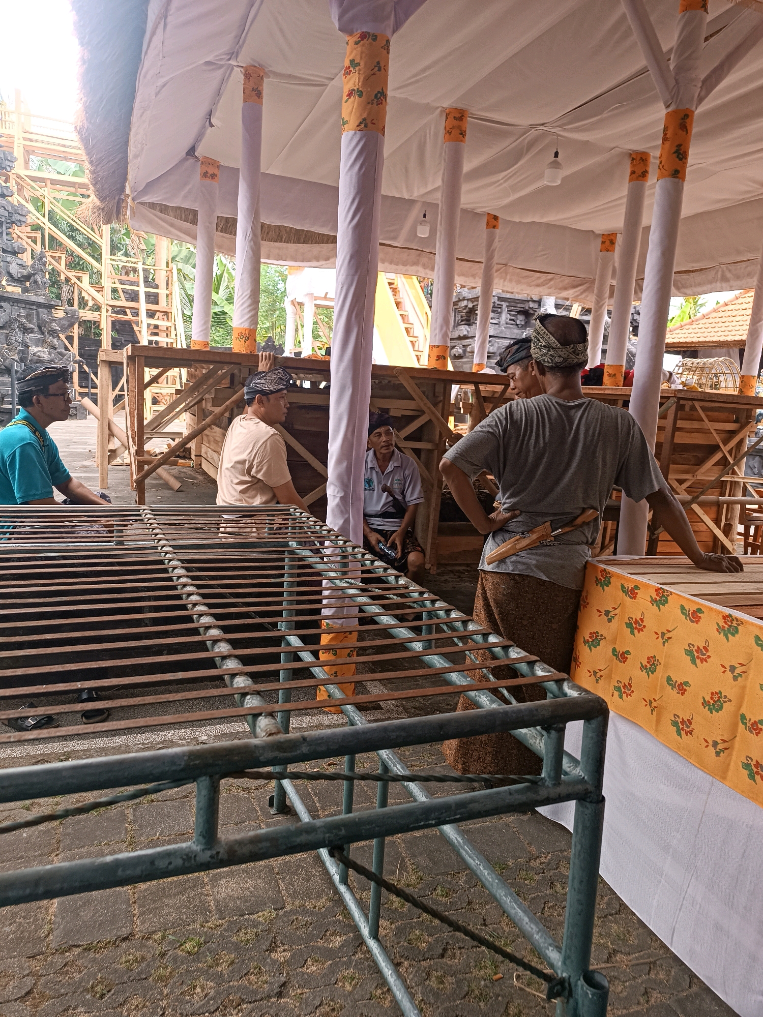The image depicts an outdoor scene under a canopy supported by white columns, likely in the city of Selemadeg, Indonesia. Four men are visible. In the foreground, a metal grate structure runs horizontally across the frame. To the left, a man in a teal shirt and glasses is seated, facing left. Next to him, a man in a light brown shirt is also seated, facing left. Behind them, a wooden structure with stairs is visible. To the right, a man wearing a grey t-shirt and a brown patterned sarong stands with his back to the camera, his left hand on his hip and his right arm resting on a table covered with a yellow cloth adorned with red and green floral patterns. A knife with a wooden sheath is tucked into his sarong. Another man in a light blue shirt is seated to the far right, looking towards the standing man. The environment appears to be a marketplace or a ceremonial gathering area, with the wooden structures suggesting temporary stalls or platforms. The canopy is white and draped, with orange and patterned fabric accents on the columns. The ground is paved with interlocking stones.