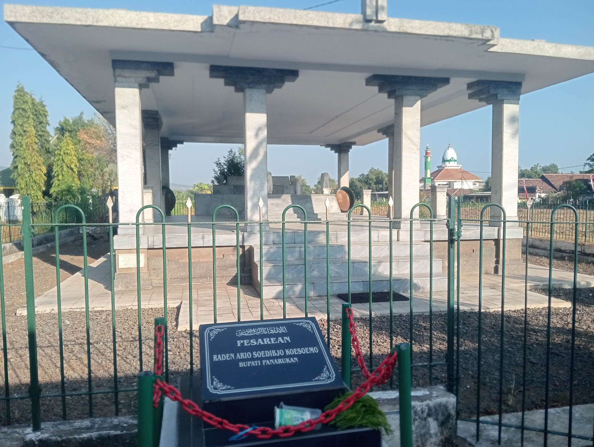The image shows a monument with a layered, flat roof supported by marble-clad pillars. The monument appears to be a tomb or memorial, with stairs leading up to a raised platform within. In the foreground, a green metal fence encloses the area, with a dark plaque resting on a base behind the fence. The plaque has white text, some of which is legible and appears to be in Arabic script above the word "PESAREAN" and below that, "RADEN ARIO SOEDIBJO KOESOEMO BUPATI PANARUKAN". A red chain is draped over the fence posts and the plaque's base. Beyond the fence, the ground is covered in gravel, and the monument itself is made of stone and marble. In the background, there are trees, a village with tiled roofs, and a mosque with a green minaret and white dome. The sky is clear and blue. The monument is identified as the Pesarean Raden Ario Soedibjo Koesoemo, located in Bloro, Indonesia.