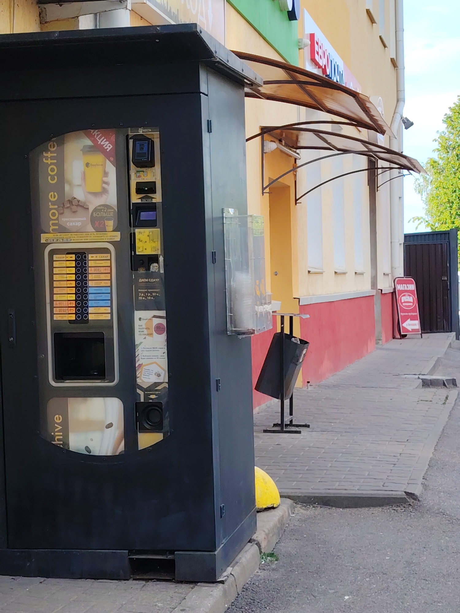 An outdoor scene in Minsk, Belarus, features a large black vending machine standing prominently on a paved sidewalk next to a building. The machine is branded with "more coffee" and "beehive" logos and displays an advertisement for a coffee promotion ("АКЦИЯ") showing a yellow cup and text about "тростниковый сахар" (cane sugar) and "X2 БОЛЬШЕ" (x2 more), suggesting a larger cup size.
The vending machine's control panel includes numerous buttons for drink selection, labeled "САХАР" (sugar) and various numbered options. Below the money slot, text indicates "ДАЕМ СДАЧУ" (We give change) in denominations of "2 р., 1 р., 50 к., 20 к., 10 к." (Rubles and Kopeks). A poster on the machine outlines "КАК ПОЛУЧИТЬ" (How to get) discounts, with an image of a loyalty card offering "50%".
The adjacent building features a yellow upper facade with white vertical stripes and a red lower portion. Several beige, translucent awnings extend from the building over the sidewalk. A partially visible sign at the top appears to read "ЕВРОПОЧТА". Further down the sidewalk, a black metal bin is positioned next to a clear plastic dispenser for cups. A red A-frame sign with "МАГАЗИН" (Shop) and an arrow points towards a dark gate. The sidewalk is paved with grey interlocking bricks, and a yellow bollard is visible at the base of the vending machine. The sky is clear, indicating daytime.