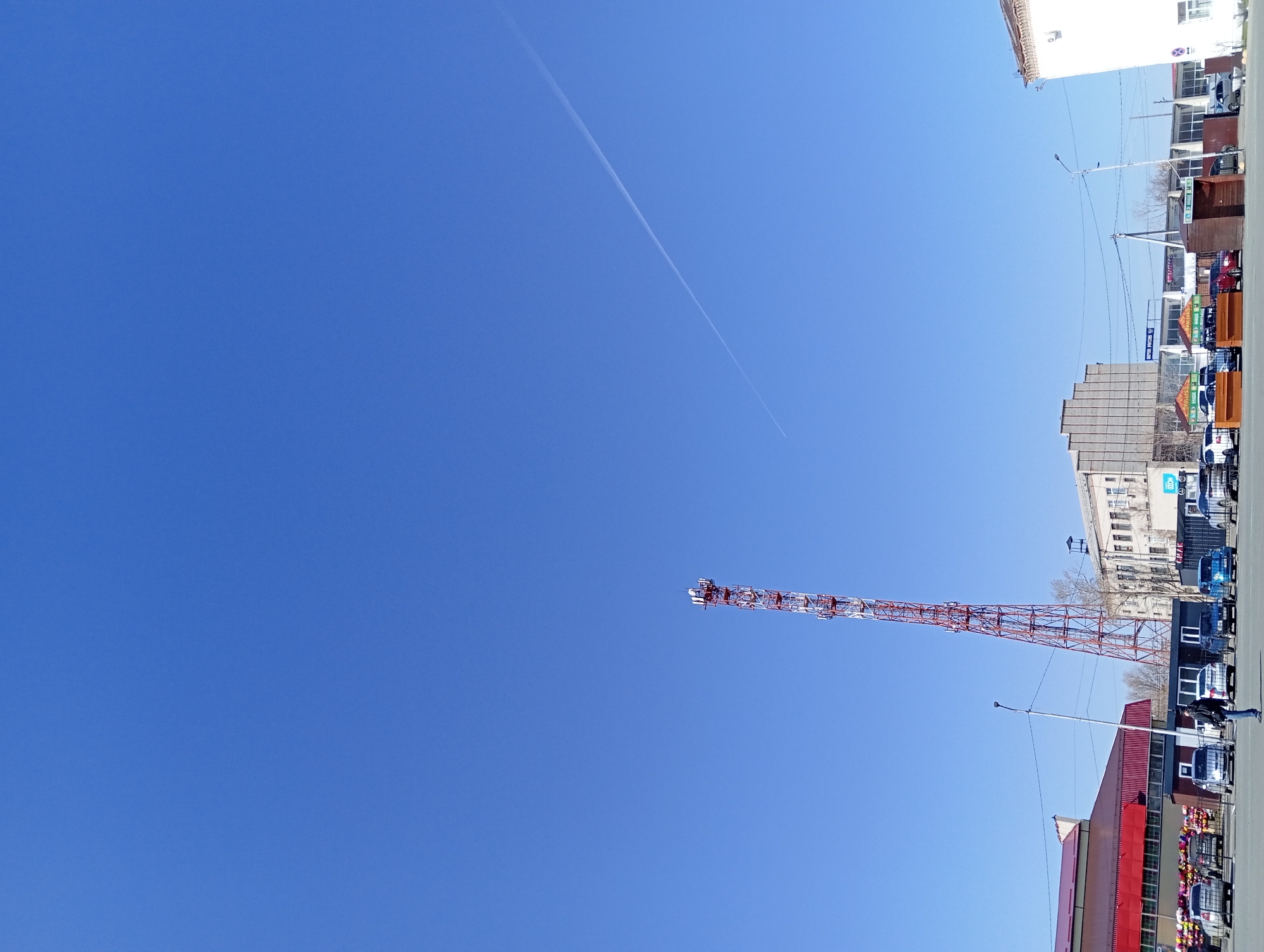 The image depicts a clear, blue sky above a street scene in Yurgamysh, Russia. A contrail from an aircraft is visible in the upper portion of the sky. A tall, red and white telecommunications tower stands prominently in the middle ground. Adjacent to the tower are various buildings, including a white multi-story structure, a red and tan building, and other commercial establishments. Automobiles are parked along the side of the road. A few pedestrians can be seen walking. A utility pole with a street lamp is also visible.