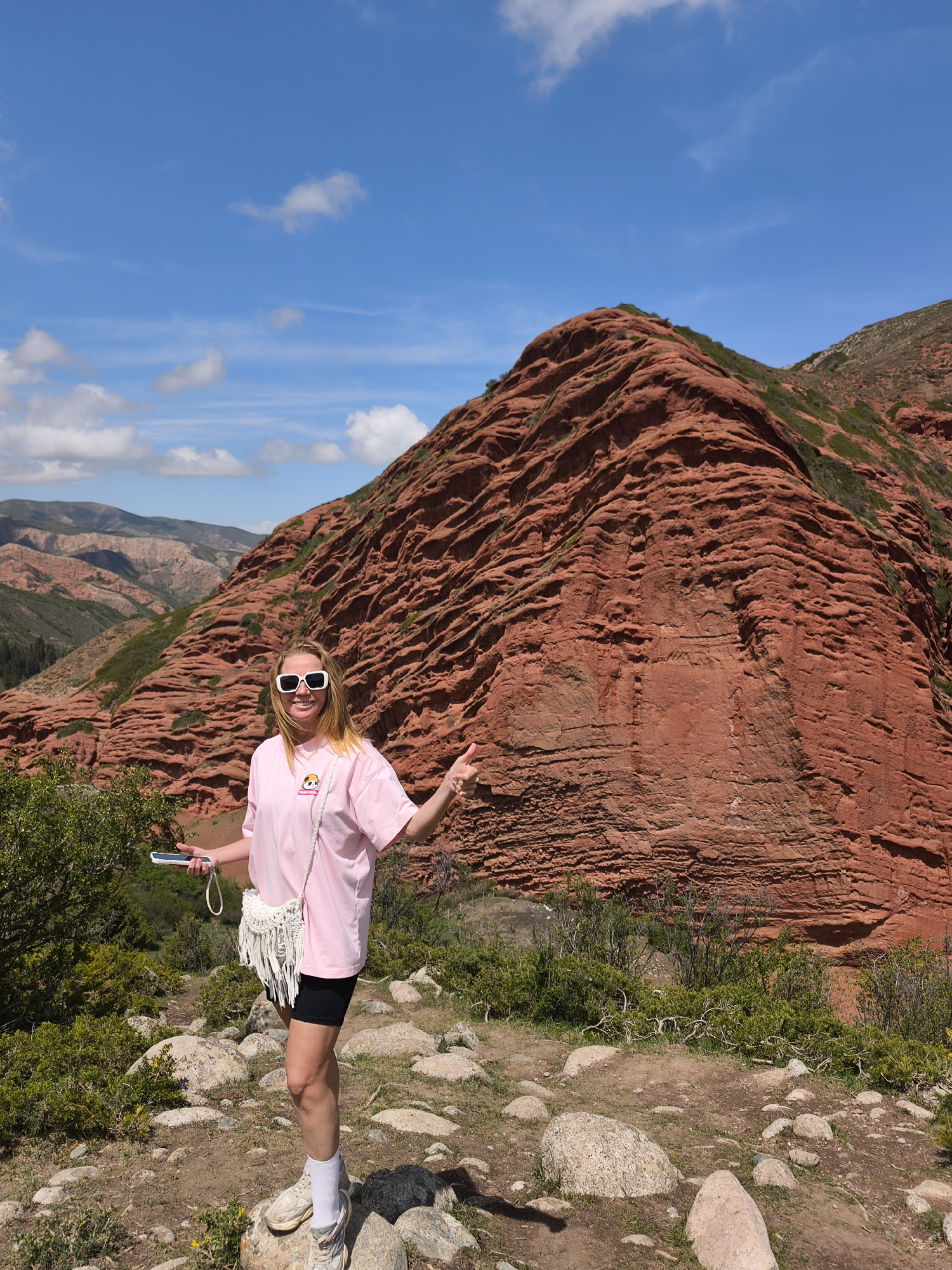 This outdoor photograph captures a woman posing in front of striking red rock formations in Jeti-Ögüz, Kyrgyzstan, on a sunny day. The scene features a clear blue sky with scattered white clouds. The woman, with blonde hair, is wearing white sunglasses, a light pink t-shirt with a small cartoon graphic on the front, black shorts, white socks, and light-colored sneakers. She carries a white woven fringed cross-body bag and holds a smartphone in her left hand. She is smiling broadly and giving a thumbs-up gesture with her right hand, indicating enjoyment or approval of her surroundings.
Behind her, a massive red rock mountain dominates the frame, displaying intricate horizontal layering and eroded textures. The ground where she stands is a dirt path with various sized rocks and sparse green shrubbery. In the distant background, a valley extends with more undulating reddish and green hills under the bright daylight, consistent with the mountainous landscape of Jeti-Ögüz. The vibrant red hue of the rocks contrasts with the green vegetation and blue sky, highlighting the unique geology of the region. The overall mood of the image is bright and cheerful, reflecting an enjoyable outdoor adventure. No discernible text is clearly visible on any objects.