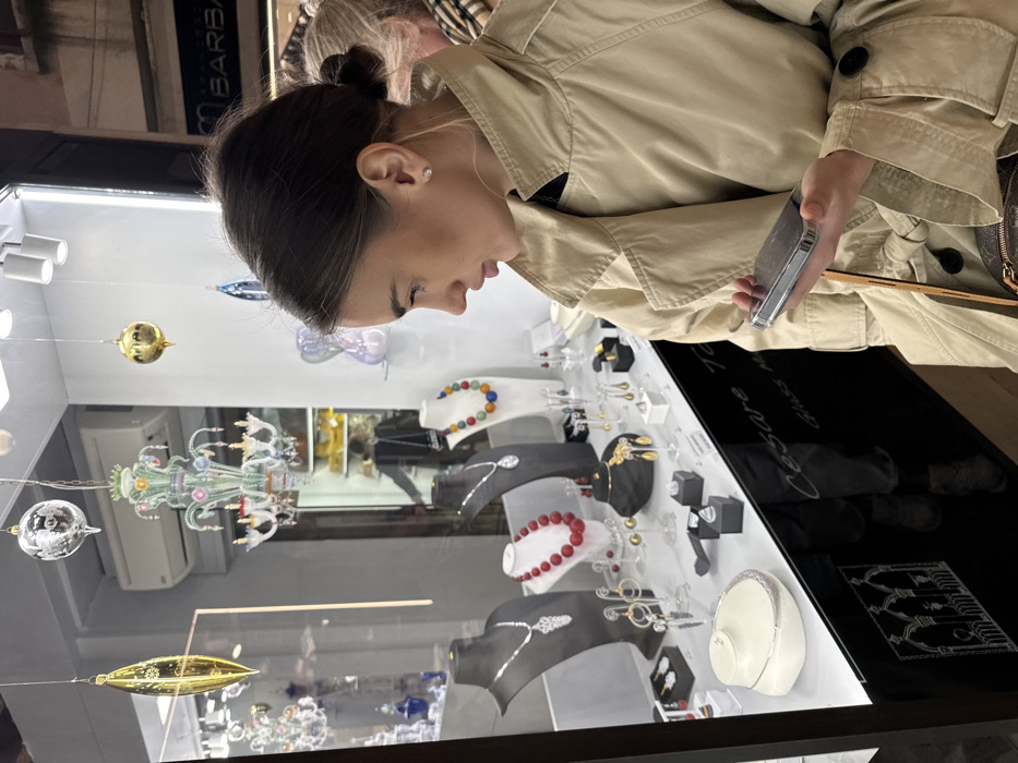 A young adult female stands facing left, intently observing a retail shop window in Venice, Italy. She wears a light beige trench coat over a dark garment and has brown hair styled in a bun, with a small stud earring visible in her right ear. Her left hand holds a silver-cased smartphone horizontally below chest level. A brown shoulder bag strap, possibly from a monogrammed purse, is visible on her right side.
The brightly illuminated shop window displays a variety of jewelry and glass art. Multiple necklaces are presented on black and white busts, including silver-toned pendants, a red beaded necklace, and a multi-colored beaded necklace. Various rings and earrings are arranged on stands and in small boxes. Several ornate glass ornaments, including a gold elongated form, a silver mirrored sphere, a gold sphere, and a multi-colored glass chandelier, hang from the display ceiling. The lower portion of the window glass shows white text reading "Cesare Tagliapietra" and "Glass Master," alongside a white outline depicting Venetian Gothic arches.
Reflected in the upper right section of the window is a partial sign for "BARBARO." Inside the store, beyond the window display, an individual in a dark jacket is reflected, along with shelves containing yellow-packaged items. A second person with light hair, wearing a patterned scarf, is partially visible behind the primary subject, also looking towards the shop window.