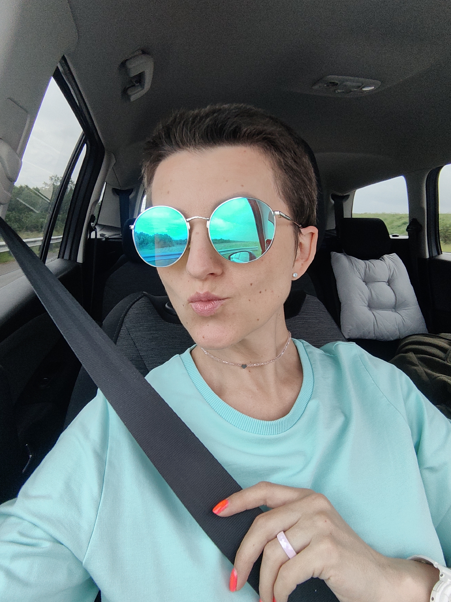 A woman with short brown hair and light skin is sitting in the passenger seat of a car, wearing round, reflective sunglasses, a turquoise sweatshirt, and a thin necklace. Her nails are painted bright orange. A pink and white ring is on her right ring finger. A black seatbelt is visible across her chest. The interior of the car is dark, with a grey seat and headrest visible behind her. A light grey tufted cushion is on the back seat. The background through the car window shows a cloudy sky and greenery. The image was taken in the vicinity of Дзямідавіцкі сельскі Савет, Belarus.