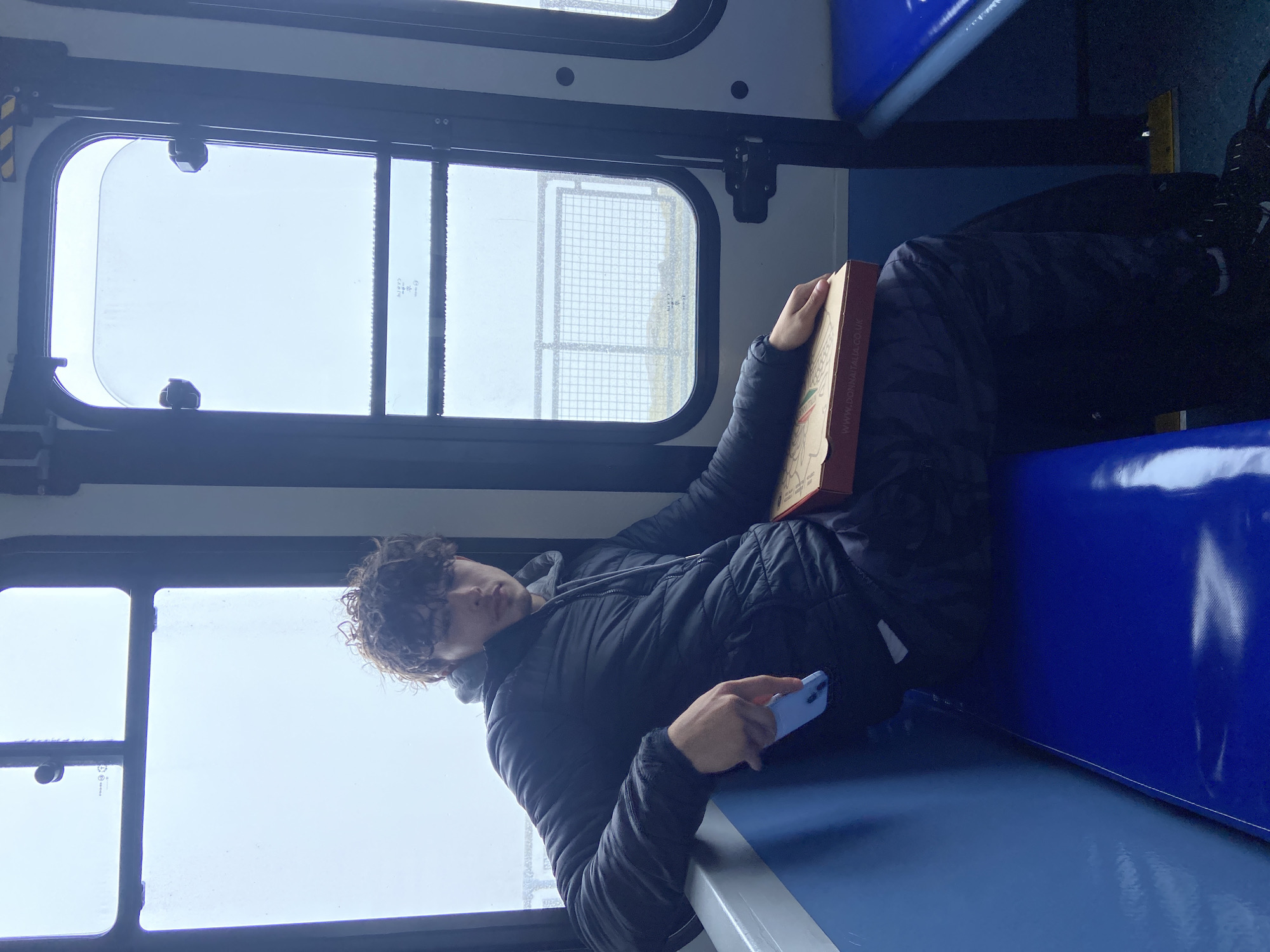 A young man lies on a blue bus seat in Caernarfon, United Kingdom, holding a phone in his right hand and a pizza box with the website "www.donnaitalia.co.uk" visible, propped up against his torso. He wears a black puffer jacket and dark trousers, his feet visible on the right side. The bus windows show an overcast sky and structures including a chain-link fence. The bus interior is marked by blue seating and a white wall with black window frames.
