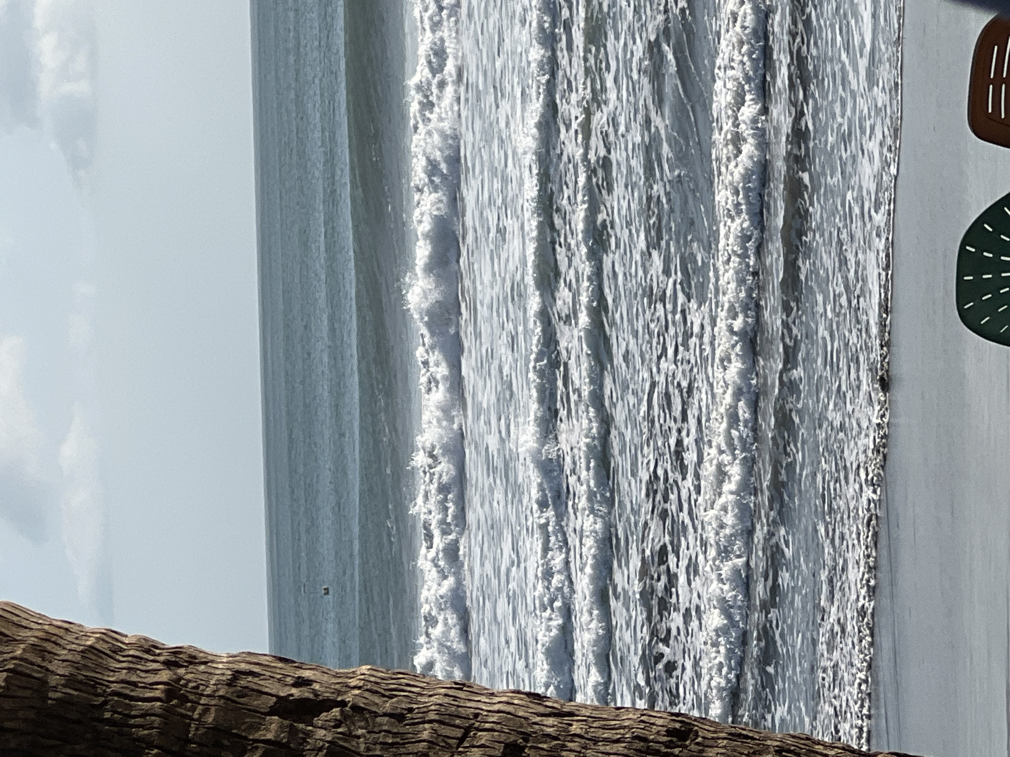 A rough-textured palm tree trunk occupies the left foreground of the image, obscuring the left edge of the scene. Beyond the tree trunk, a wide expanse of the ocean stretches horizontally across the frame. Multiple waves are breaking and rolling towards a sandy beach, creating white foam and churning water. In the far distance, a small, dark object, possibly a buoy or a small boat, is visible on the ocean's surface. The sky above is a pale blue with scattered, wispy clouds. The bottom of the image shows the edge of a sandy beach. Partially visible in the lower right corner are parts of two beach chairs, one green and one brown. The scene is indicative of a beach location in Kuta, Indonesia, known for its surfable waves and coastal scenery.