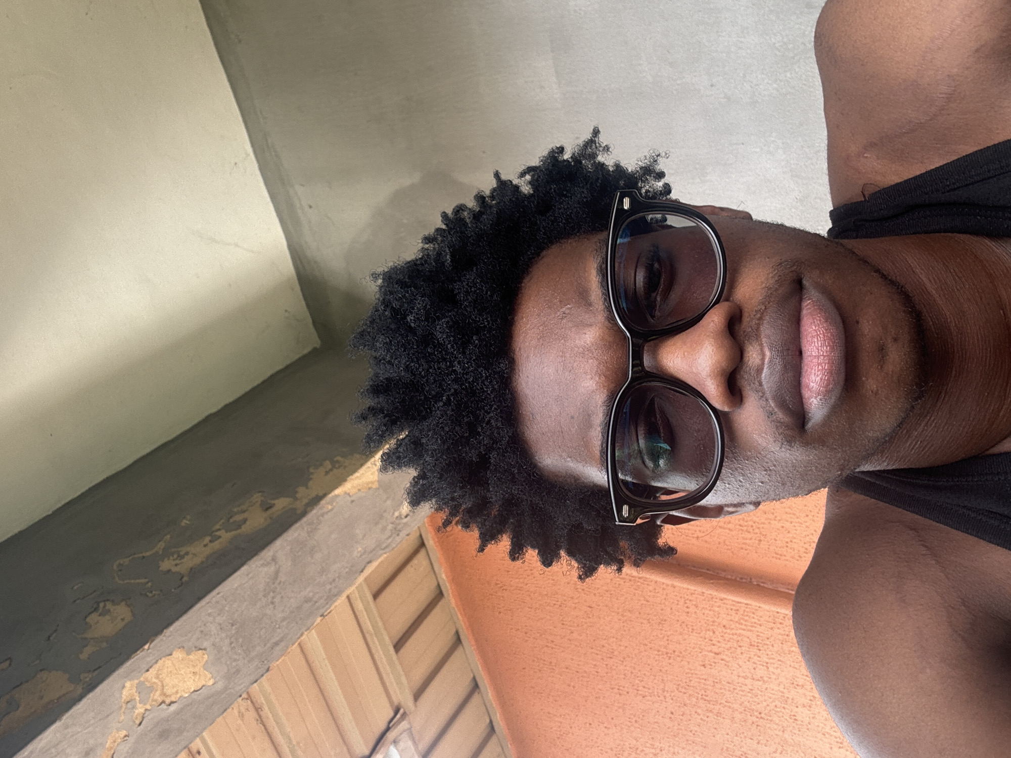 The image shows a close-up, frontal shot of a Black male with dark, curly hair and dark-rimmed glasses. He is wearing a black tank top. The background consists of an orange textured wall on the left, a wooden ceiling structure above and to the left, and a light gray concrete wall on the right. A portion of a beam with peeling paint is visible in the upper central area. The lighting suggests an outdoor or semi-outdoor setting. The overall impression is that of a casual selfie taken outdoors.