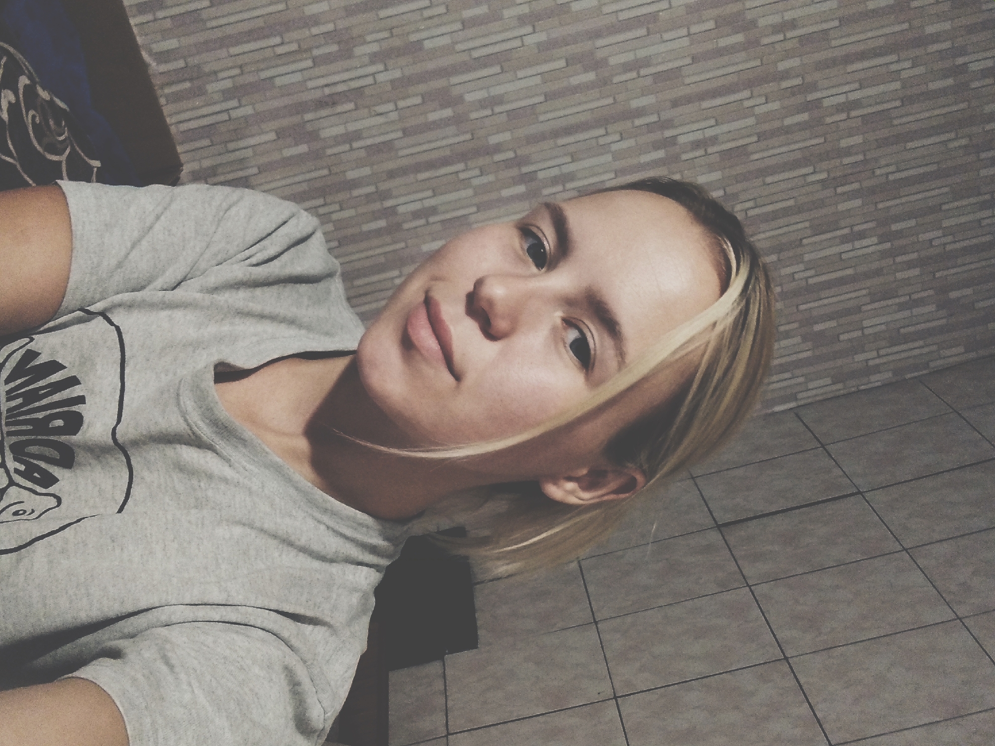 A young woman with blonde hair tied back is pictured in a selfie. She is wearing a grey t-shirt and is situated in an indoor environment. The background consists of textured walls. Based on the provided context, this scene is located in the city of Radisne, Ukraine.