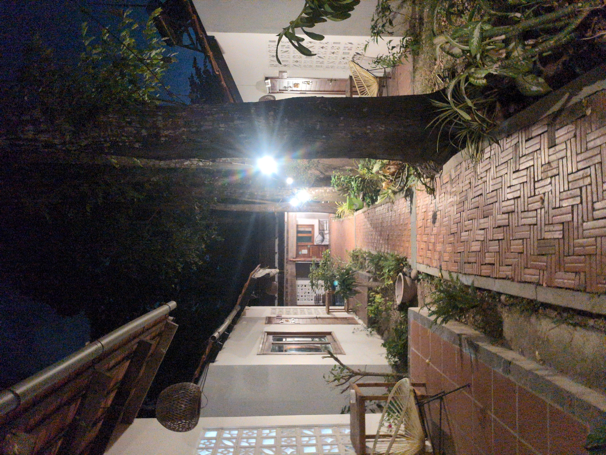 A dimly lit outdoor scene in Phú Quốc, Vietnam, shows a narrow brick-paved path extending between two buildings. String lights illuminate the path, which is partially obscured by a large tree trunk. The buildings are two stories high and constructed of white stucco. The building on the right has a brick facade and contains hanging flowerpots and potted plants. A rattan chair sits on the right side of the path. A portion of a roof and a hanging basket are visible in the lower left corner. The left side of the path is dominated by the tree and shadows, obscuring the building behind it. A small patch of blue sky is visible above the tree.