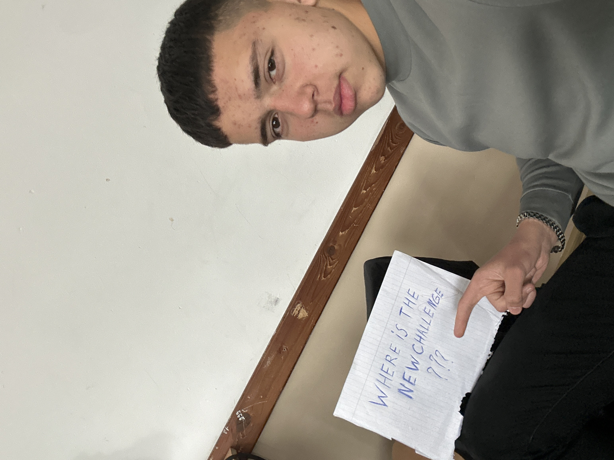 A person is holding a piece of paper that says "WHERE IS THE NEW CHALLENGE ???". The person is wearing a grey sweatshirt, black pants, and a silver bracelet. The background is a white wall with a brown wooden trim. The location is unknown and cannot be determined from the image.