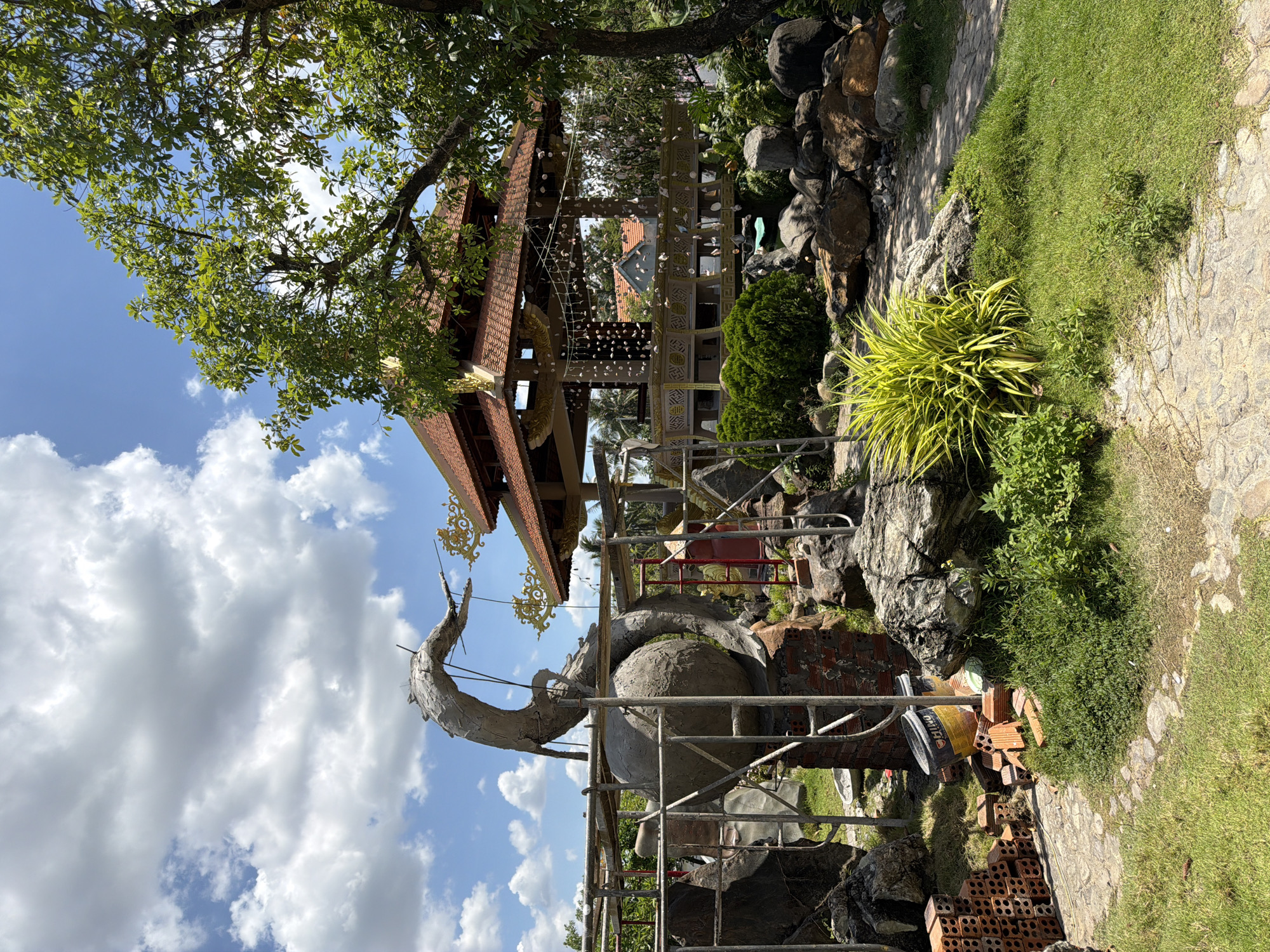 The image captures a sunny outdoor scene, likely a garden or park under construction, featuring an unfinished sculptural element and a traditional Vietnamese-style pavilion. In the foreground, a large, grey, unfinished sculpture resembling a dragon or mythical creature, coiled around a spherical base, is partially supported by scaffolding. Bricks and construction materials, including a yellow bucket, are scattered around the base of the sculpture. The ground is a mix of grass and a stone pathway. Behind the sculpture, a multi-tiered pavilion with an orange tiled roof is visible, decorated with hanging ornaments. Lush green bushes and large rocks are interspersed throughout the scene, and a large tree with dense foliage stands to the right. The sky is bright blue with scattered white clouds. The location is identified as Tuy An Nam, Vietnam.