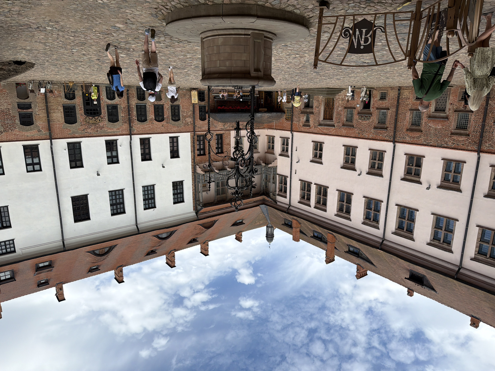 The image shows an upside-down view of a cobblestone courtyard surrounded by brick and white buildings. A stone well with a circular base is centrally located in the upper portion of the image. Several people are visible walking through the courtyard. In the foreground, two people stand near a decorative metal fence with the letters "WB" visible on it.
The scene appears to be an outdoor public space, possibly a historic town square or courtyard. The buildings have multiple windows with dark frames and a tiled roof structure is visible extending from the lower section of the buildings. The sky, with scattered white clouds, occupies the bottom part of the image, reflecting the inverted scene above. The time of day appears to be daytime, and the weather seems clear. There are no discernible emotions, but the presence of people suggests a normal day of activity.