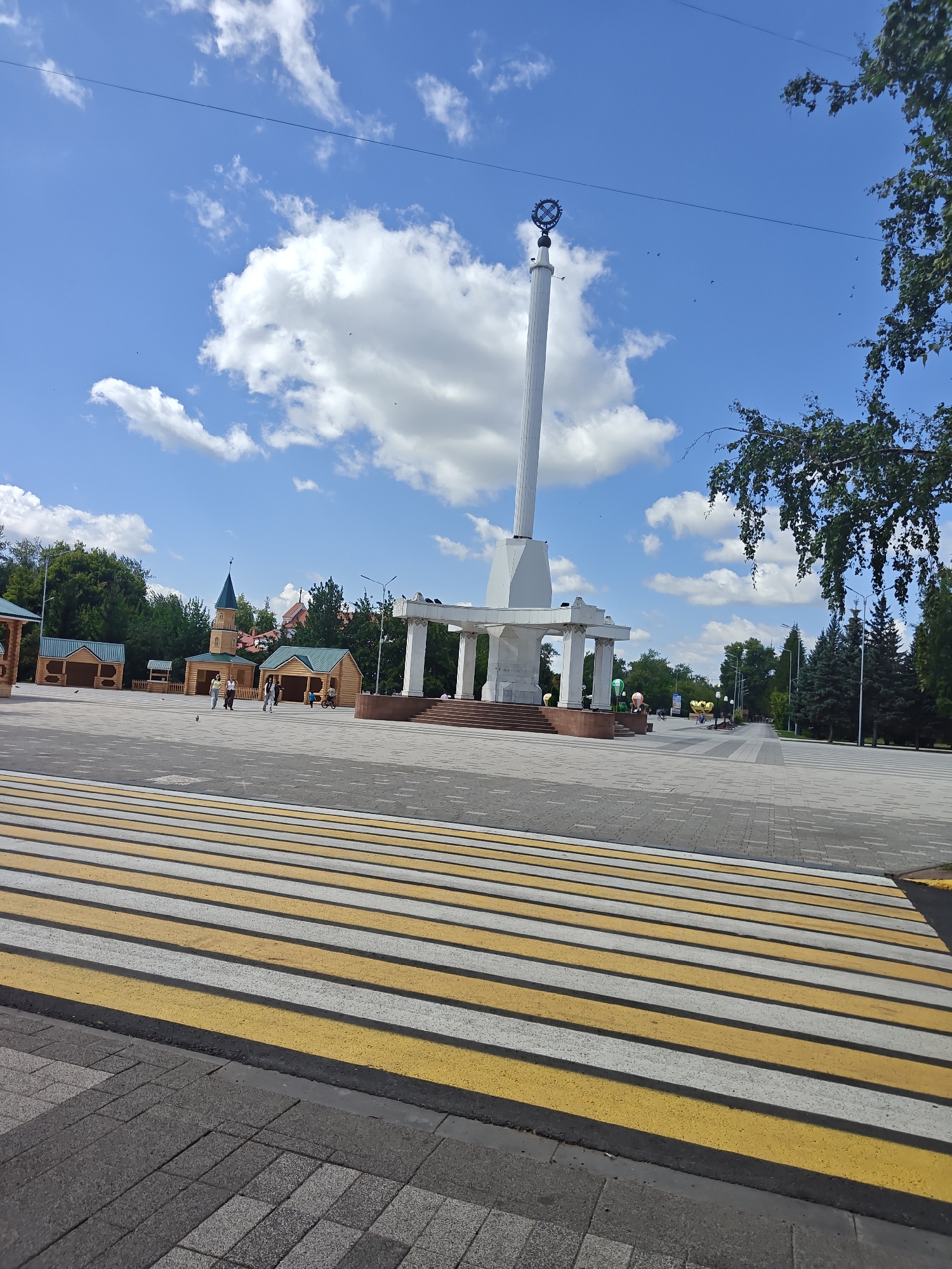 A sunny day in Petropavl, Kazakhstan, is depicted in this outdoor scene, with a prominent monument dominating the center. The monument is a tall, white, cylindrical pillar topped with a circular emblem. It stands on a raised platform with white columns and a staircase leading up to it. In the foreground, a pedestrian crossing with yellow and white stripes is visible on a paved walkway.
To the left of the monument, there are several small wooden structures, one of which resembles a mosque with a green spire. People are walking and standing in this area. Behind these structures, a dense line of green trees forms the background, with some buildings peeking through.
To the right of the monument, the paved walkway continues, lined with more trees, including tall fir trees. The sky is a vibrant blue, dotted with fluffy white clouds. A few birds are visible in the sky. A large tree with green leaves hangs into the frame from the upper right corner. The overall impression is of a well-maintained public space on a clear day.