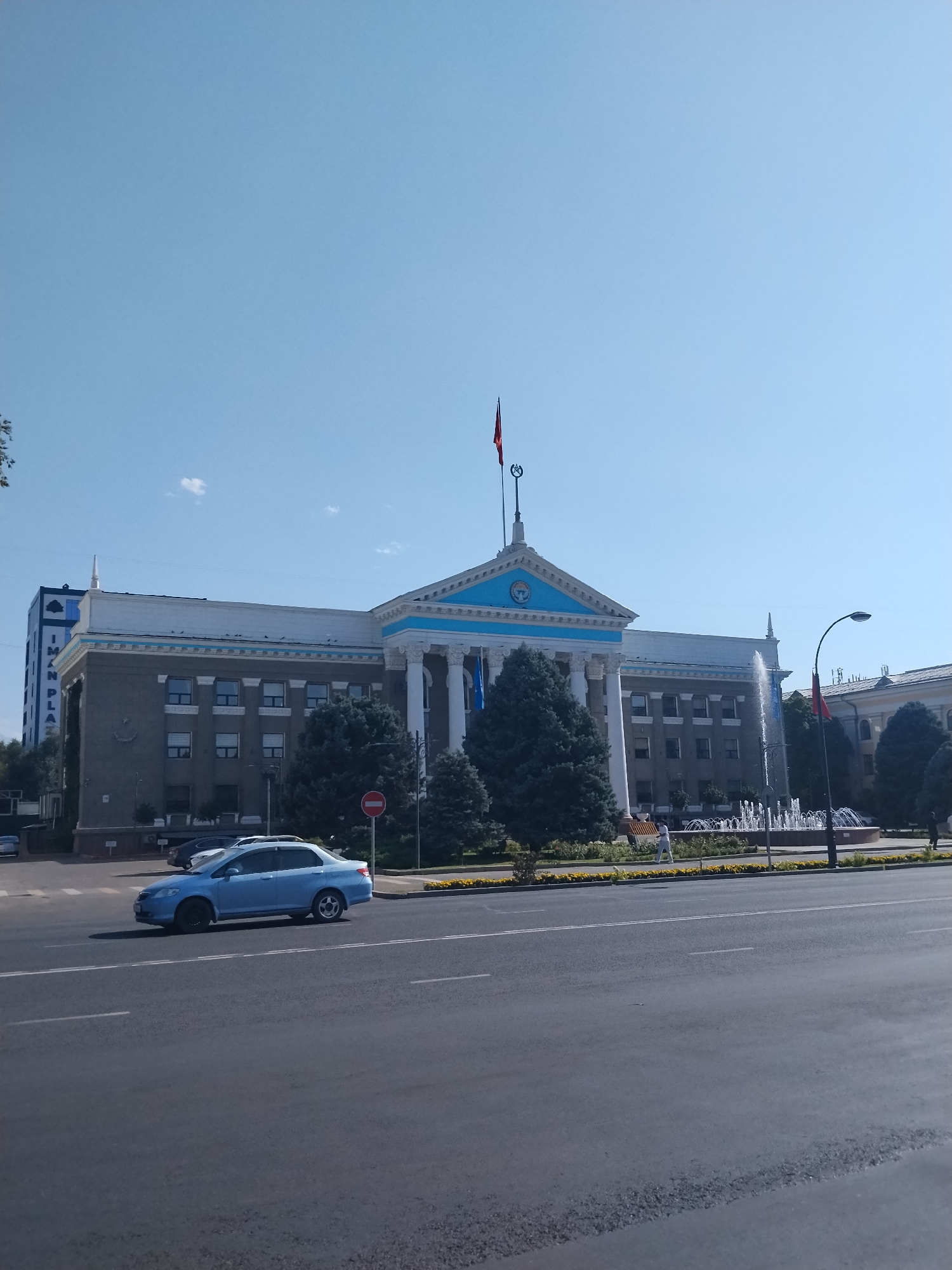 The image captures a daytime scene in Bishkek, Kyrgyzstan, focusing on a prominent building with classical architectural elements, likely a government or administrative structure. The building is light gray with white columns supporting a blue-accented pediment. A flag, red with a yellow emblem, flies from a pole atop the structure. In front of the building, a multi-tiered fountain is active, with water spraying upwards. Several evergreen trees are visible in front of and around the building. The foreground shows a paved road with a light blue sedan driving on it. A red circular sign with a white horizontal bar, indicating "no entry," is positioned on the sidewalk to the left of the fountain. The sky is clear and blue. To the far left, a modern tall building with blue accents can be seen.