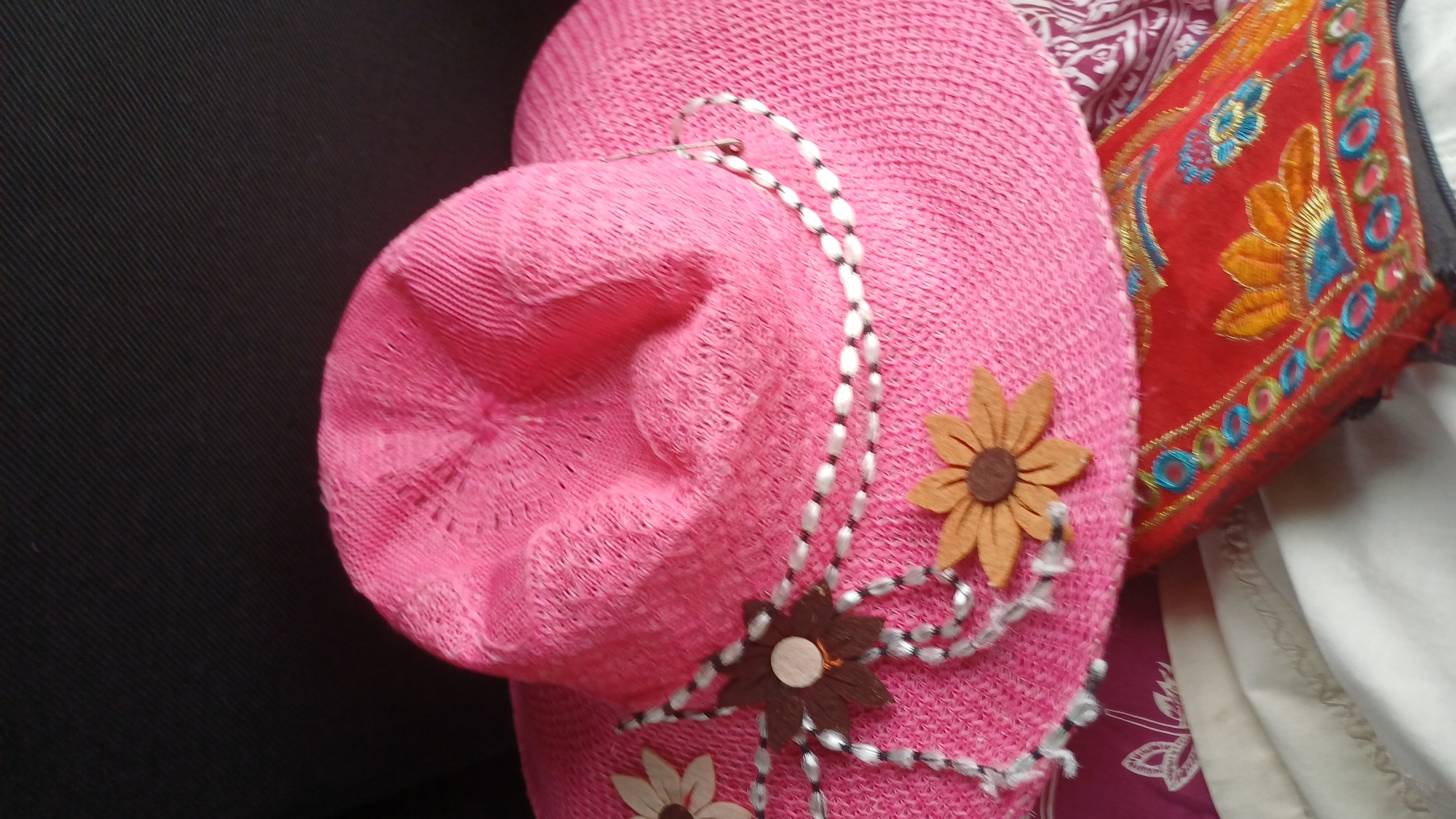 A bright pink woven sun hat with a wide brim is lying on top of a folded red fabric item with embroidered floral designs. The hat is decorated with two felt flowers, one light brown and one tan, each with a brown center. A beaded string with white and black beads, along with some white fringe, is wrapped around the hat's crown and attached to the flowers. The background is dark and textured, appearing to be a dark fabric. The bottom right portion of the image shows a patterned fabric, possibly a blanket or tablecloth, with white and maroon designs. There are no people or visible actions taking place in the image. The scene is likely indoors, given the lighting and background. The location of this image in the city of Киясово, Russia, is not visually discernible from the image itself.