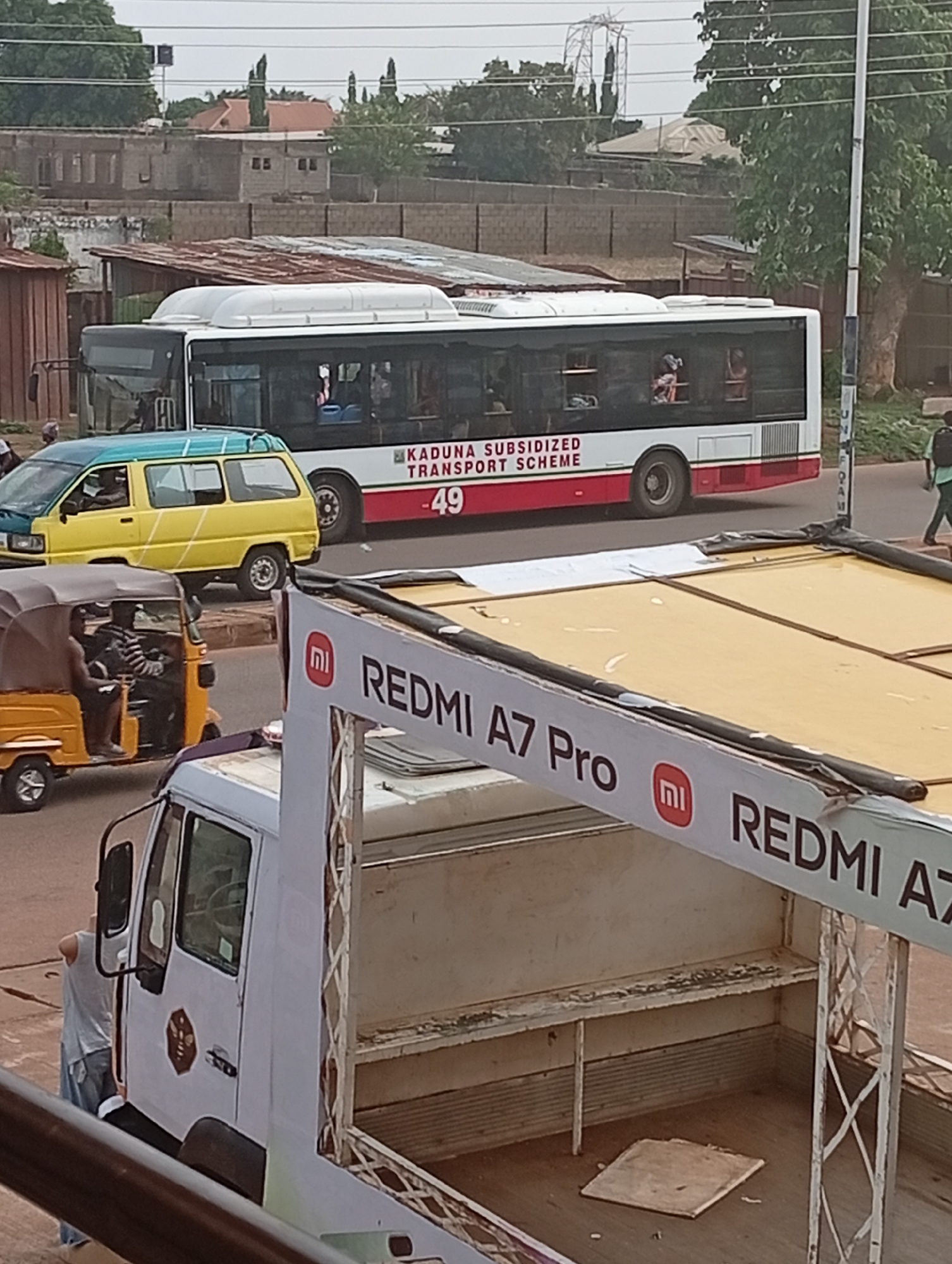 The image depicts a street scene in what appears to be Sabon Tasha, Nigeria. Prominently featured is a white bus with red accents labeled "KADUNA SUBSIDIZED TRANSPORT SCHEME - 49". Beside it is a yellow and blue small van. In the foreground, there is a white truck, bearing the advertisement "Redmi A7 Pro". An auto-rickshaw and a person are positioned adjacent to the truck. Other pedestrians are seen on the sides of the road. Buildings and foliage are present in the background.