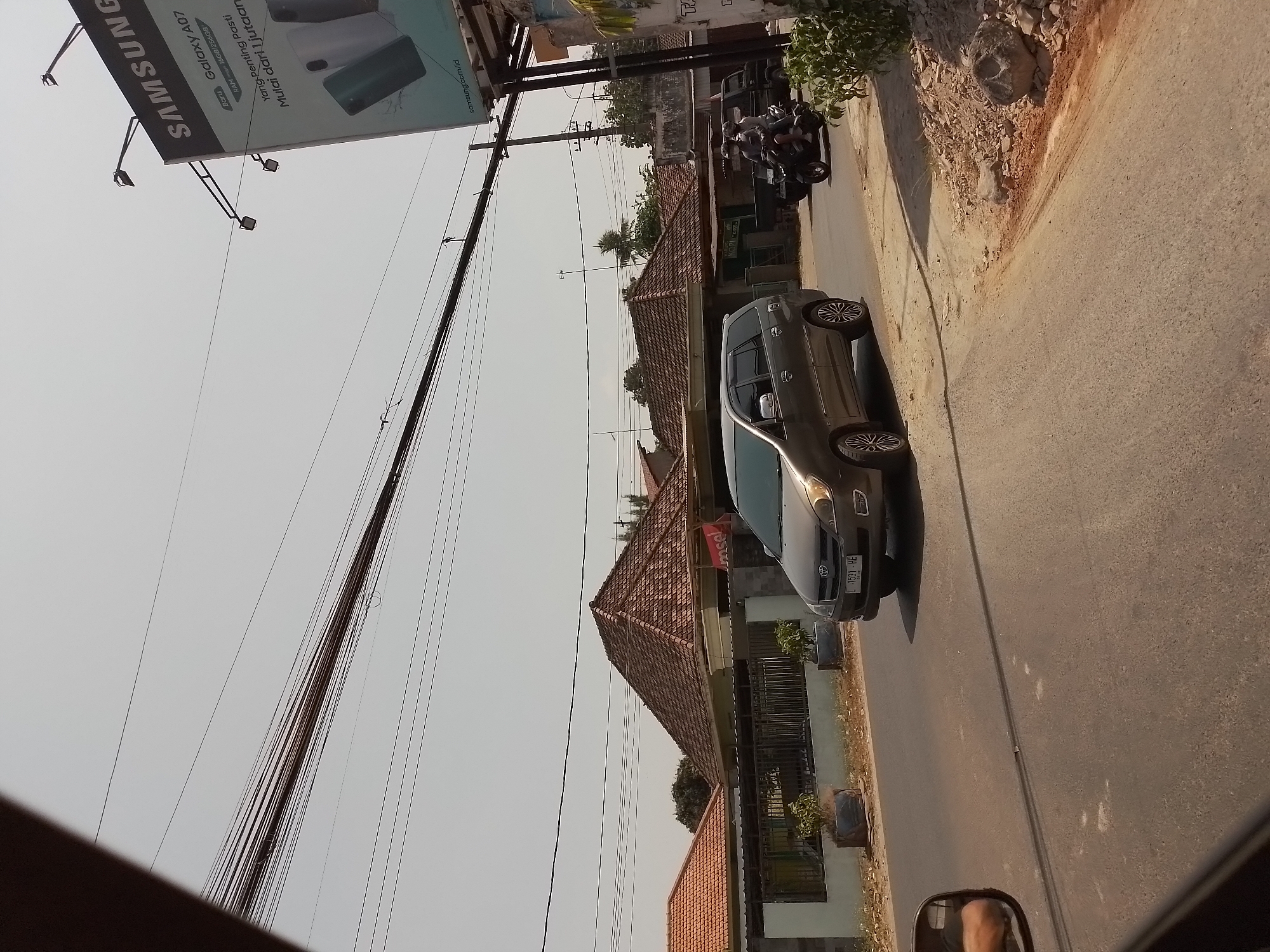 The image captures a street scene in Wates, Indonesia, on a bright, sunny day. The foreground shows the asphalt road and the side mirror of a vehicle, reflecting a portion of the interior. A dark grey minivan, identified by its license plate "L 1531 HE," is traveling on the road. Further down the street, a black motorcycle with two occupants is also moving.
On the left side of the road, a residential area is visible, characterized by houses with tiled, pyramid-shaped roofs and white fences. Several potted plants are placed along the sidewalk.
Dominating the upper right portion of the image is a large Samsung billboard advertising the Galaxy A07. The billboard is supported by metal poles and illuminated by spotlights. Overhead, a dense network of power lines crisscrosses the sky. The environment appears somewhat dry, with patches of dirt and loose rocks visible on the roadside.