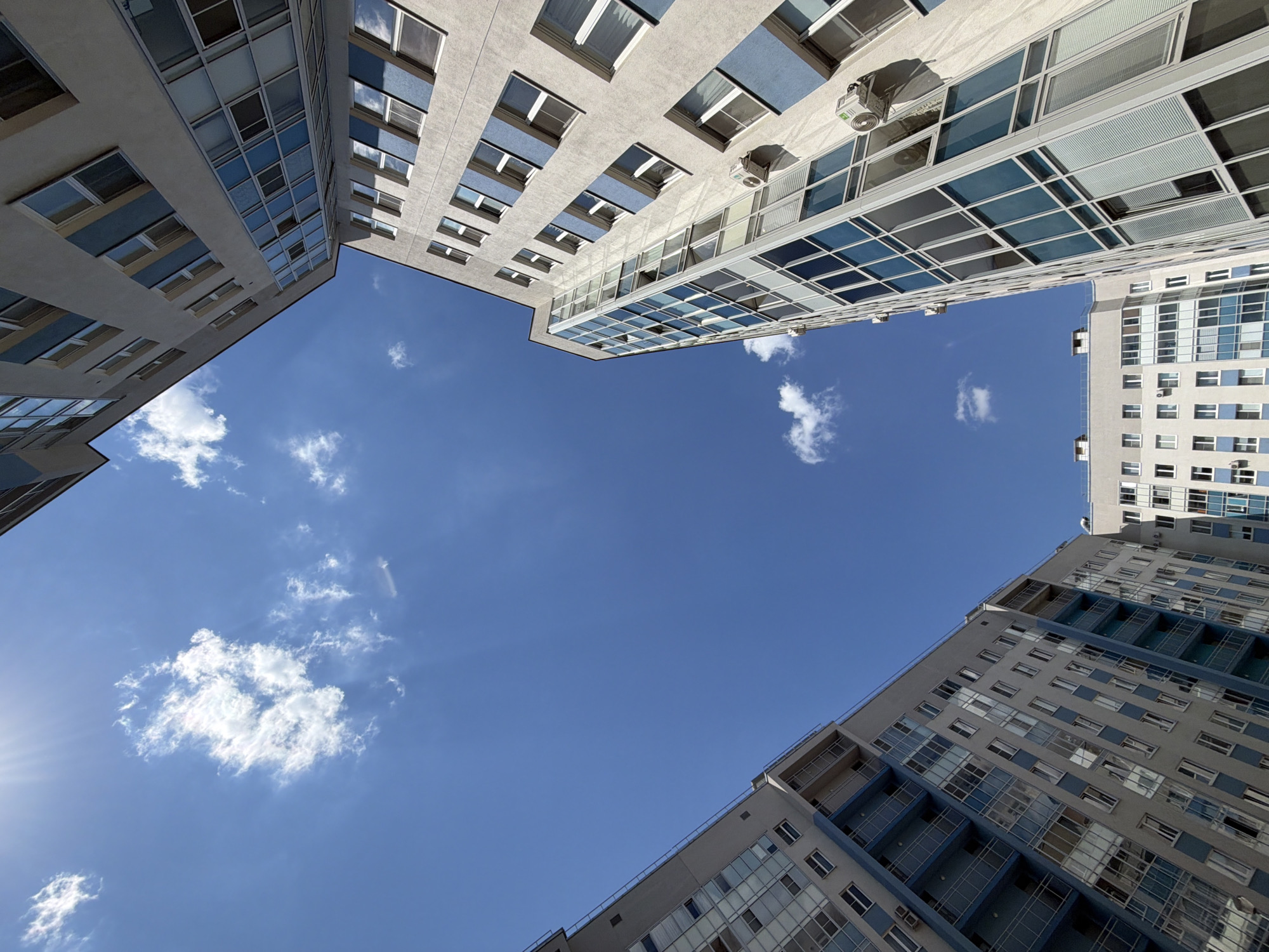 The image captures an upward-looking view of a vibrant blue sky framed by the modern facades of several high-rise buildings. The perspective creates an urban canyon effect, with the towering structures defining the edges of the frame.
The buildings are predominantly light-colored, featuring numerous windows that are either reflective, mirroring the sky, or show internal blinds. Some sections of the architecture include blue paneling or trim, and several external air conditioning units are visible on the building surfaces. The architectural style appears contemporary, characteristic of modern city developments.
The majority of the central image is occupied by a clear, bright blue sky dotted with scattered, fluffy white clouds. The sun is partially visible in the bottom-left corner, casting a bright lens flare and indicating a sunny day.
There are no people or specific activities taking place; the scene is a static architectural and sky shot. The bright sun and clear sky suggest it is daytime, likely mid-morning to afternoon, with pleasant weather conditions. This urban environment is located in Saint Petersburg, Russia. No visible text is present in the image.