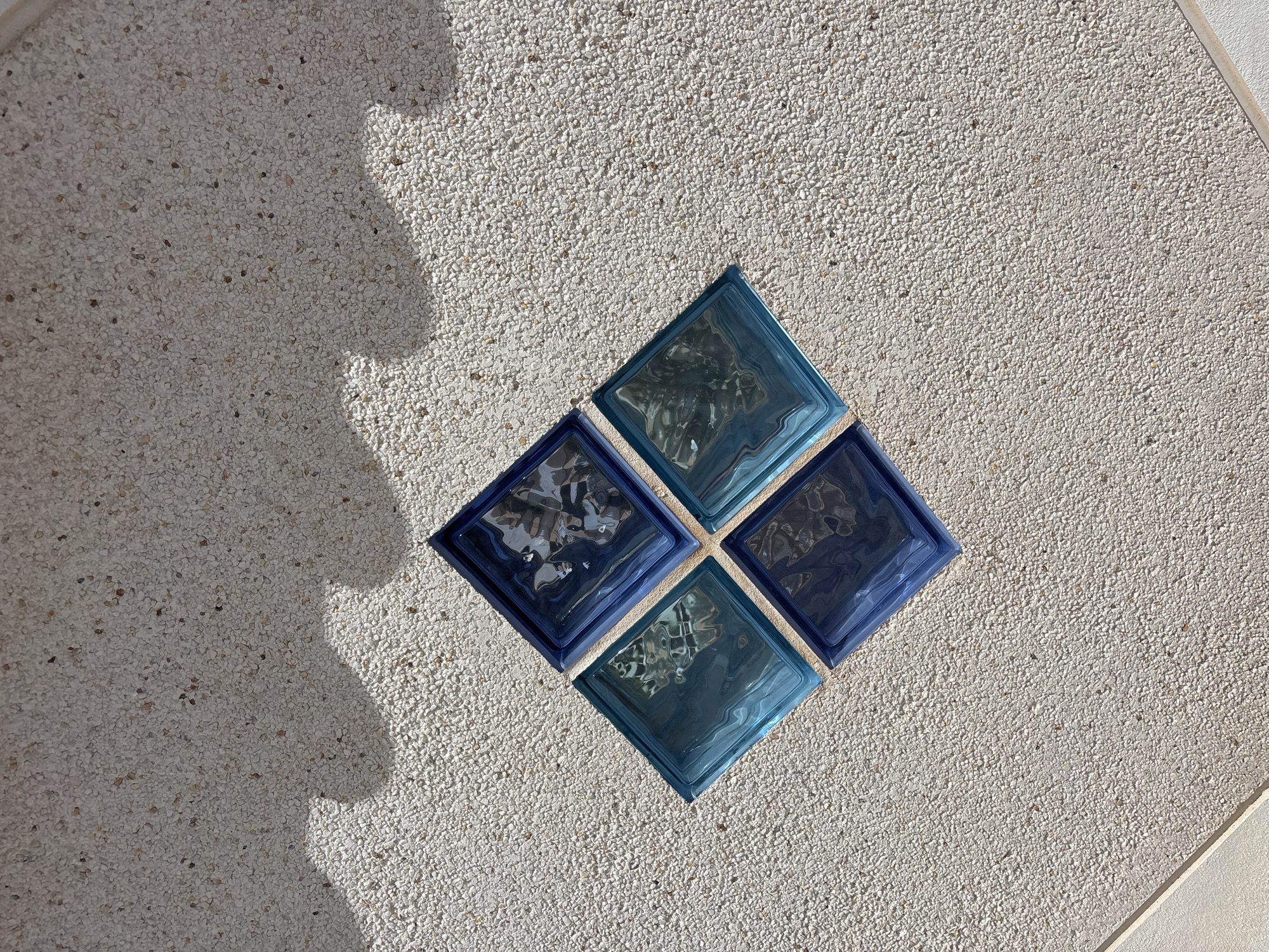 This is a close-up, overhead shot of a decorative wall in Orihuela, Spain. The wall's surface is a light-colored aggregate with small, varied-colored pebbles. Embedded within the wall are four translucent, blue glass blocks arranged in a diamond pattern. Two blocks are a darker, navy blue, and two are a lighter, teal blue. The glass blocks have a textured, rippled surface that distorts the light passing through them, creating interesting reflections and refractions.
To the left of the glass blocks, a shadow is cast on the wall, suggesting it is outdoors during daylight. The shadow's soft edges and the bright illumination imply it's a sunny day, possibly late morning or early afternoon. The concrete aggregate texture and the presence of the glass blocks suggest an outdoor setting, perhaps a garden wall, a patio, or a pool area. There is no text visible in the image. The overall mood is serene and visually interesting due to the interplay of light, shadow, and the textured materials.