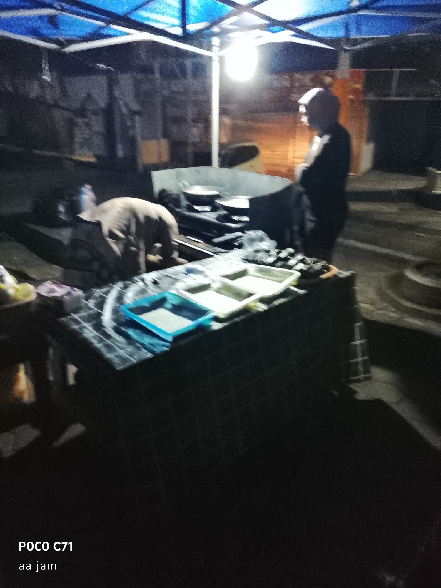 The image depicts a nighttime outdoor scene under a blue tarp shelter. In the foreground, a table is covered with a checkered cloth, displaying several white trays. A person, partially visible, is bent over the table, appearing to be engaged with something on it. Further in the background, another individual wearing a cap stands with arms crossed, looking to the side. Beyond them, there's a large round object that could be a cauldron or a cooking vessel. The environment appears to be dimly lit by artificial light, with a generally dark and somewhat unkempt setting. The text overlay at the bottom indicates "POCO C71" and "aa jami", likely denoting the device used and possibly a name or tag. The location is specified as Tasikmalaya, Indonesia.