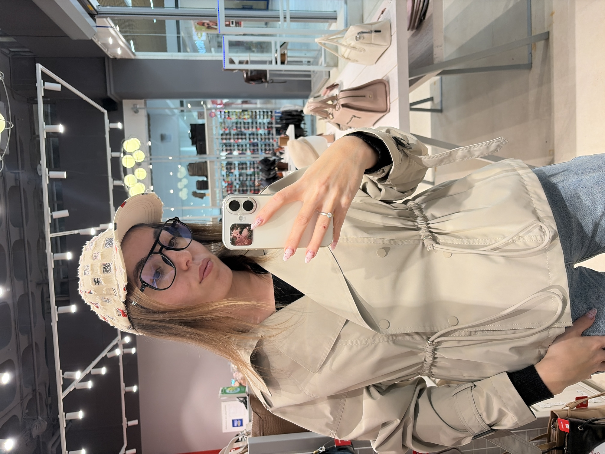 A person, possibly female, is taking a selfie in what appears to be a retail store. The individual has brown hair, is wearing glasses, a patterned hat, a beige trench coat, and jeans. They are holding a smartphone with a floral design case, presumably to capture the image. A ring is visible on their finger.
In the background, there's a display of handbags on shelves and rows of shoes. Overhead lighting is prominent, consisting of linear rows of lights. The merchandise suggests a clothing or accessories store. Based on the items visible, the location cannot be definitively identified as being in Izhevsk, Russia, or any other specific city without further contextual information.