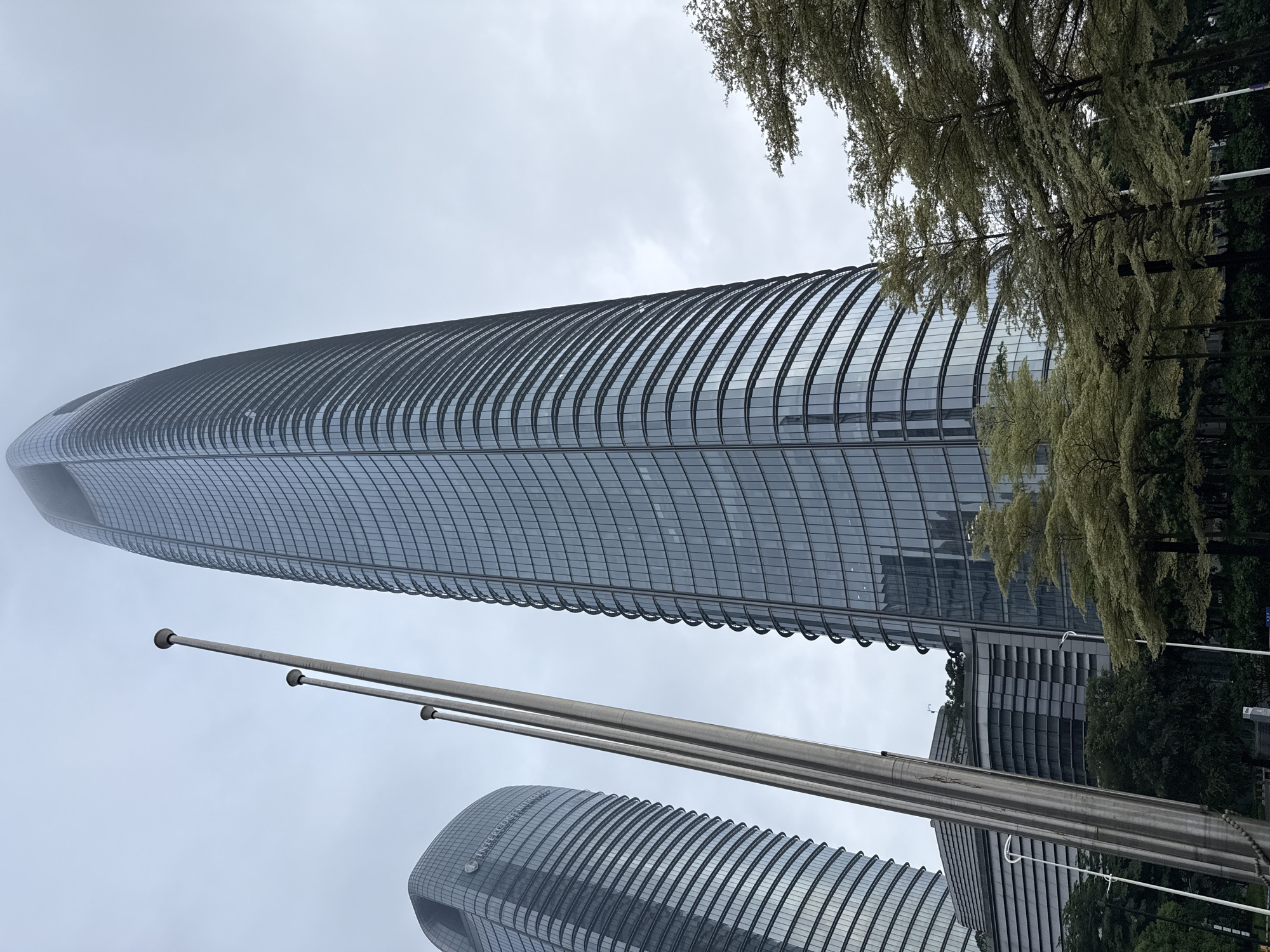 The image shows a view of the Guangzhou International Finance Center, a supertall skyscraper in Haizhu District, China. The skyscraper is a modern glass-and-steel structure with a curved facade. In front of the skyscraper, there are three tall flagpoles made of metal. The foreground of the image is dominated by trees with light-colored leaves, suggesting it is spring or autumn. The sky is overcast and cloudy. To the left of the main skyscraper, another tall building with a similar modern architectural style can be seen. The scene appears to be an urban environment, possibly a city square or park area in front of the financial center. There are no people visible in the image.