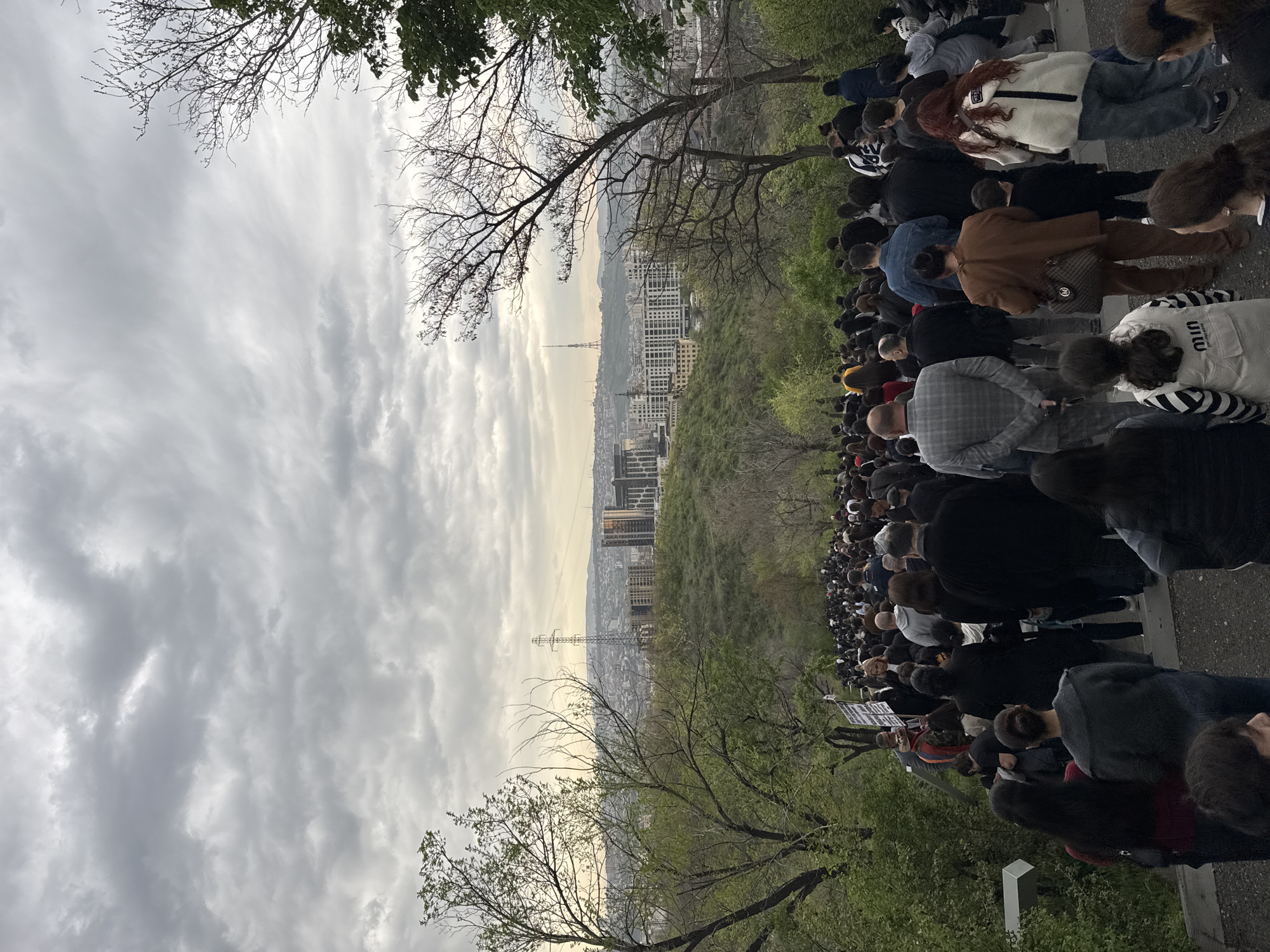 The image depicts a gathering of people on a paved area overlooking a cityscape, identifiable as Yerevan, Armenia, due to the presence of the Yerevan TV Tower visible in the background. The sky is overcast with dense cloud cover. Trees with sparse foliage frame the top of the image. The group of people appear to be mostly facing the same direction, towards the cityscape. There are individuals in a variety of clothing styles.