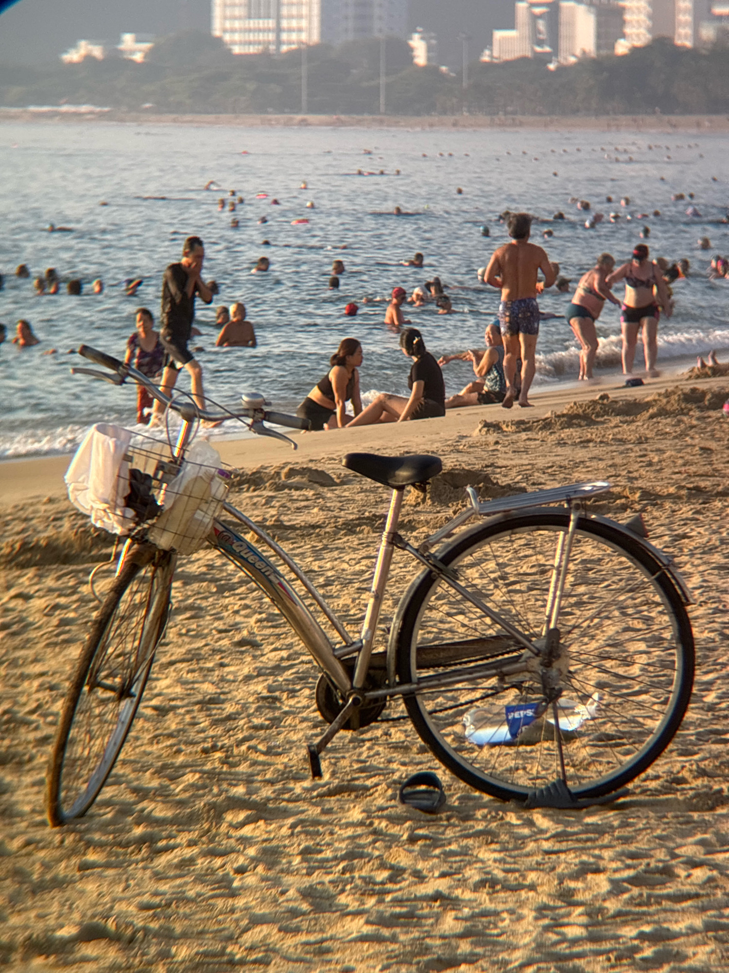 This is a photograph taken on a sandy beach in Nha Trang, Vietnam, likely during the daytime. In the foreground, a silver bicycle with a basket filled with a white cloth and some plastic bags is parked on the sand. The bicycle has a black seat and handlebars. Further back, numerous people are visible in the ocean, some swimming and others wading in the shallow water. On the shore, several individuals are sitting or standing. The background shows a hazy view of city buildings and some greenery, suggesting an urban coastal setting. The overall scene depicts a bustling beach with recreational activities taking place in the water and on the sand.
